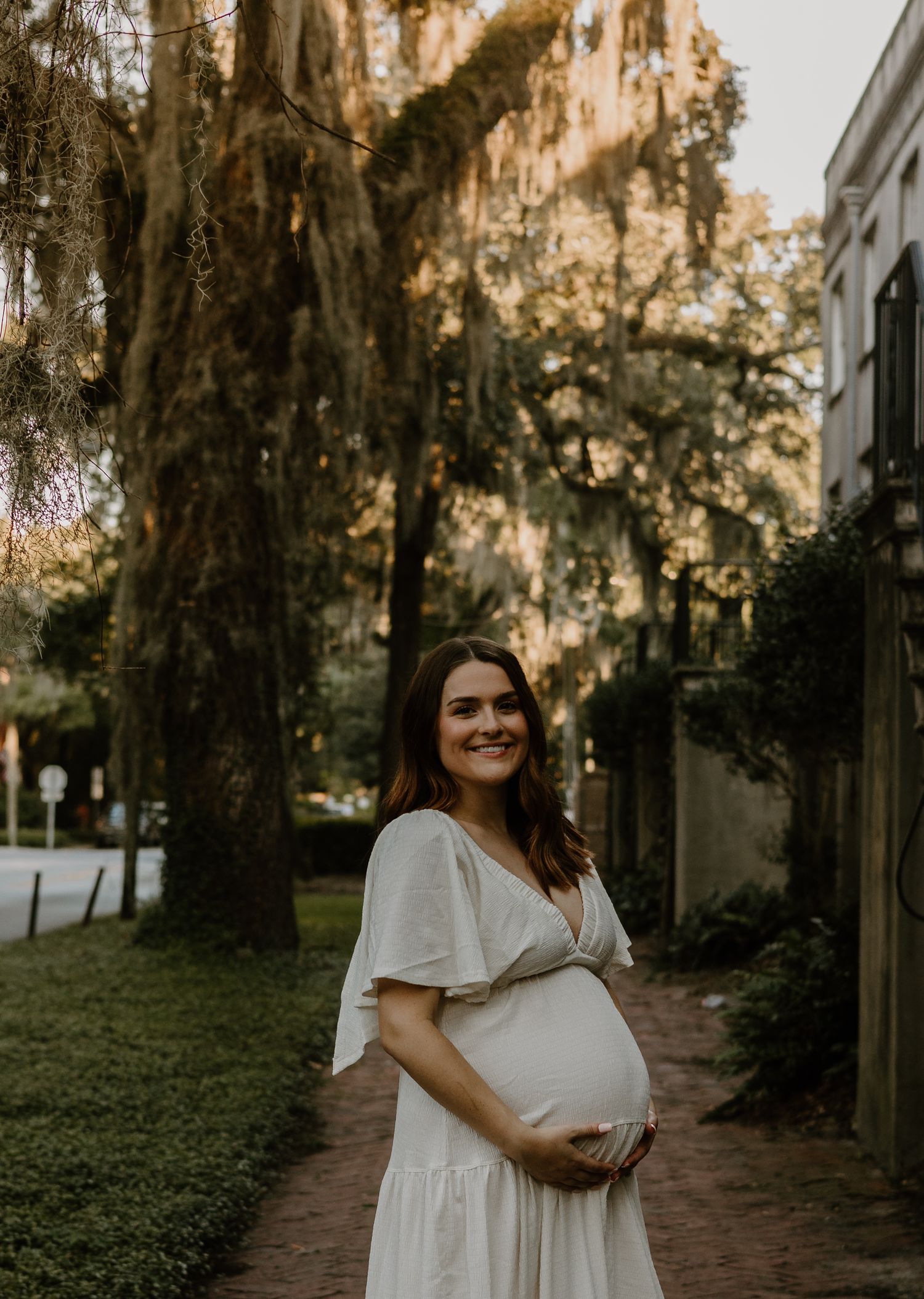 A person in a flowing white dress poses along a brick pathway with Spanish moss draped trees.