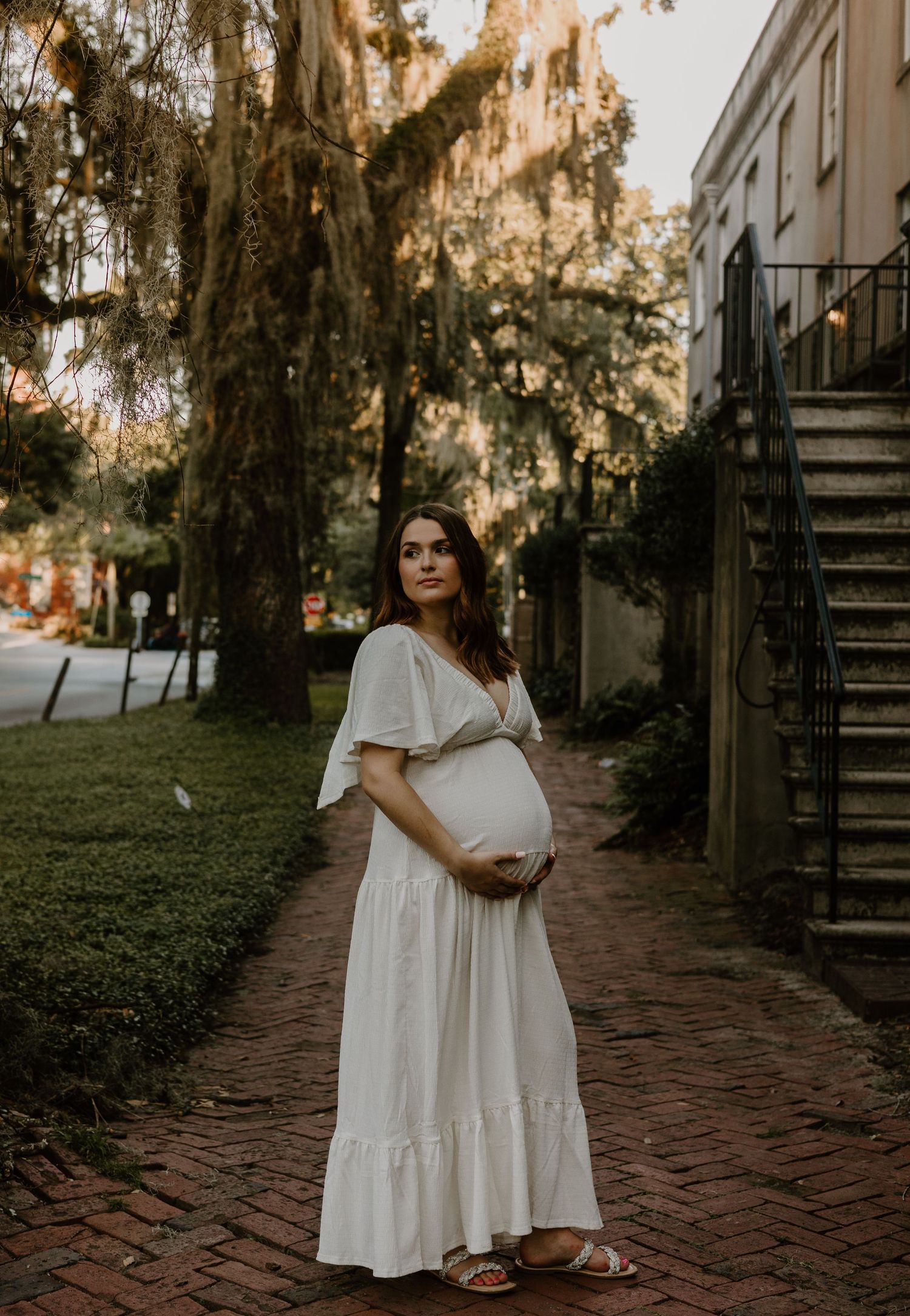 A person in a white dress poses along a brick pathway surrounded by moss-covered trees at sunset.