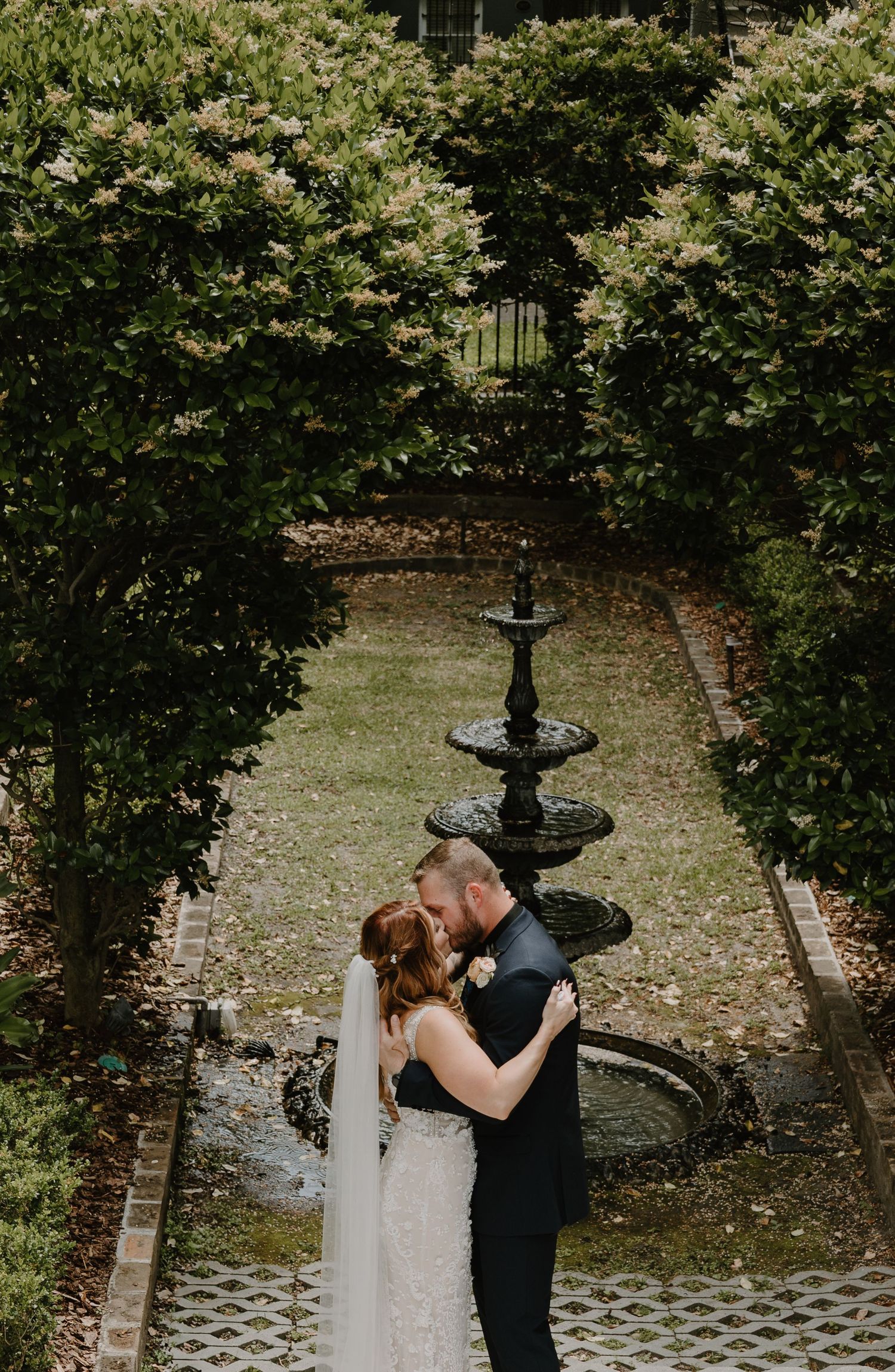 A romantic wedding moment captured in a garden setting with a stone fountain and lush greenery.