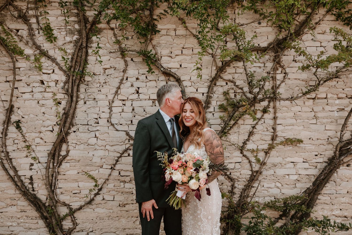 A couple poses against a white brick wall with climbing vines during a romantic photoshoot.