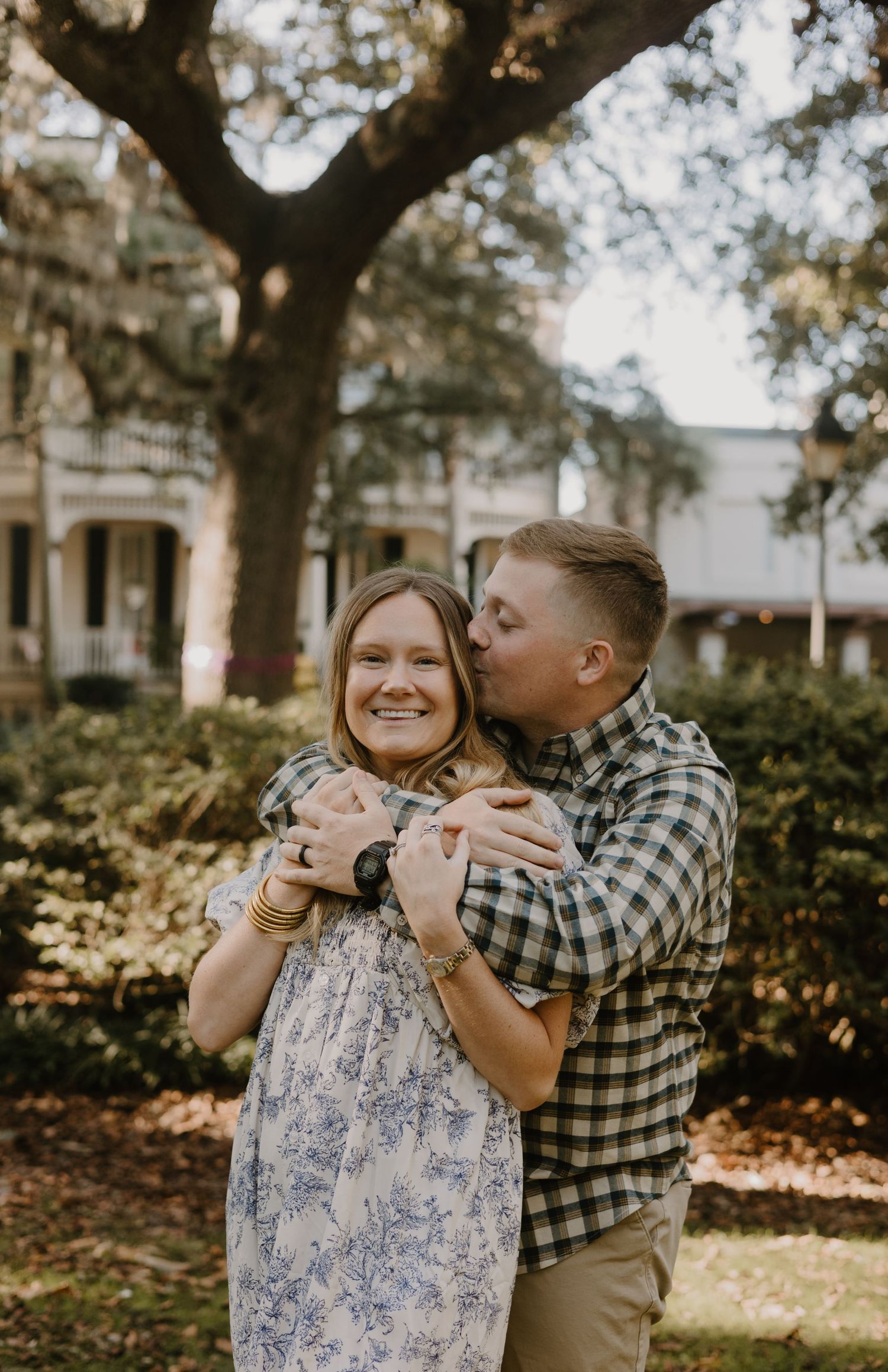 A couple embraces joyfully in a park setting with trees and buildings in the background.