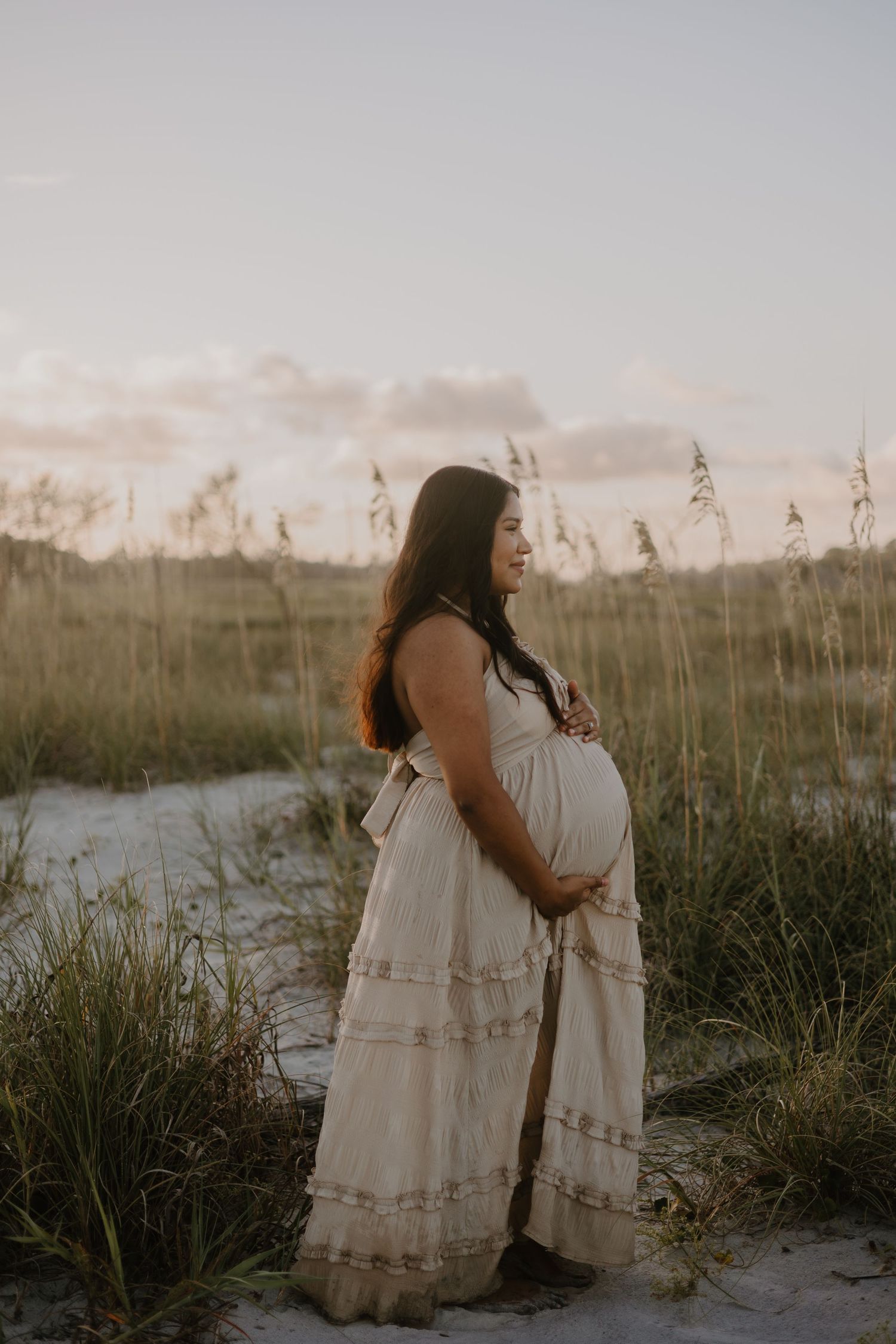 Series of dreamy beach maternity photos showing flowing white dress against coastal grasses.