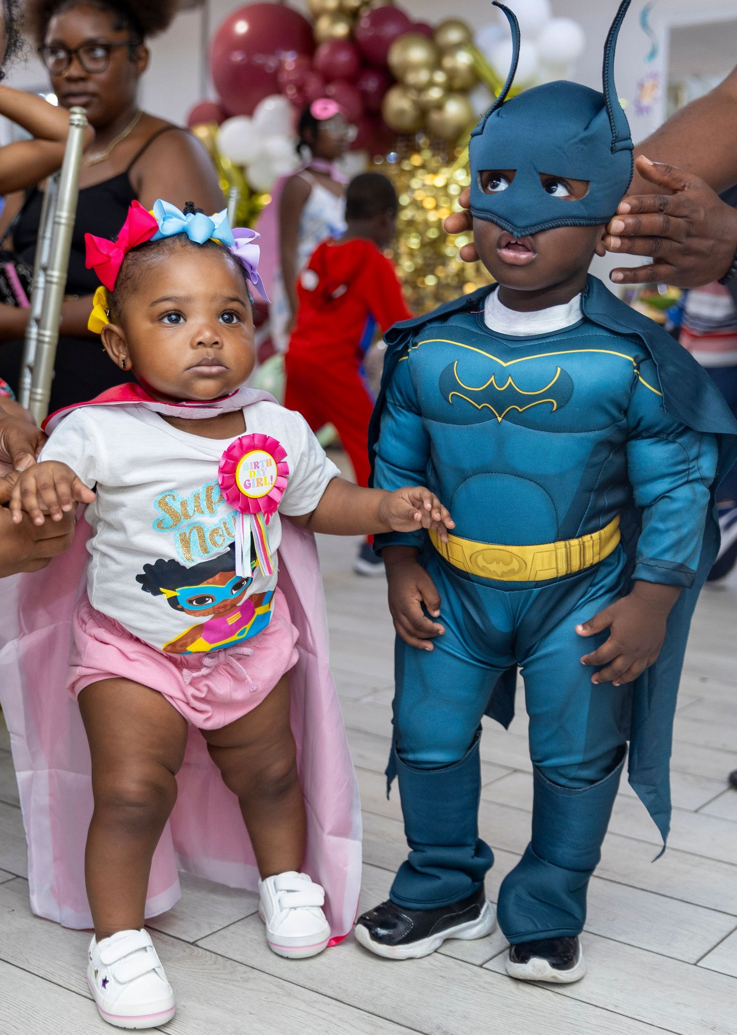 Two children at a party, one wearing a superhero costume and one in a pink outfit with bow.