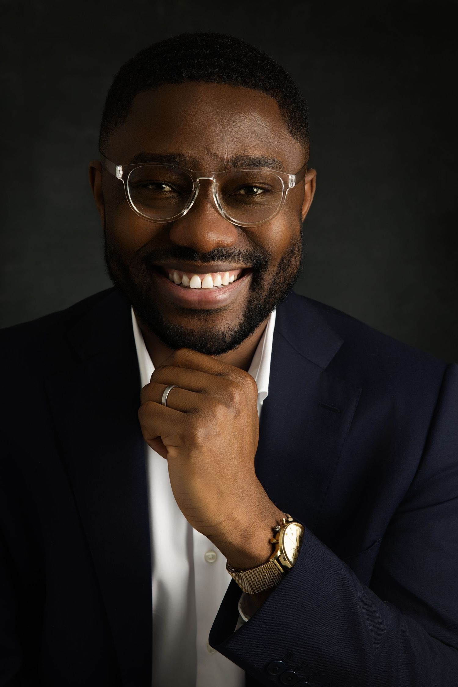 Professional headshot of someone in a navy suit and glasses against a dark background.