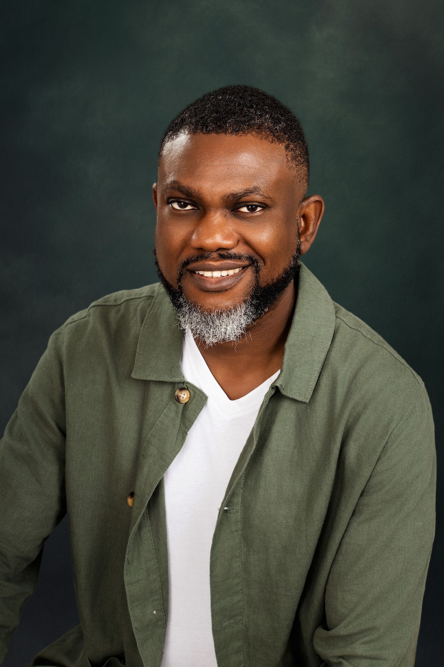 Professional headshot of someone wearing an olive green button-up shirt over a white v-neck against a dark background.