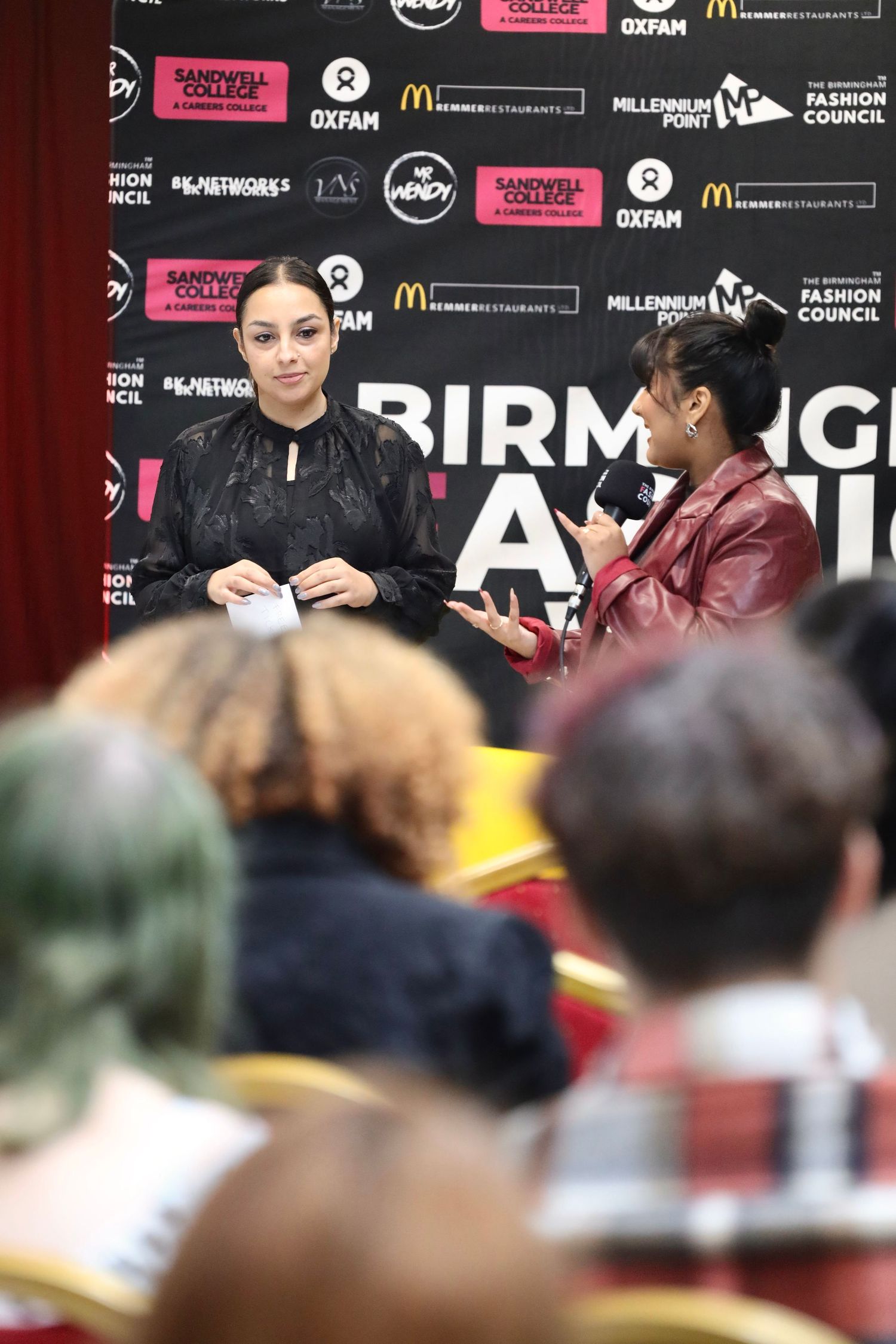 Audience members seated at Birmingham Fashion Week with event backdrop and sponsor logos visible.