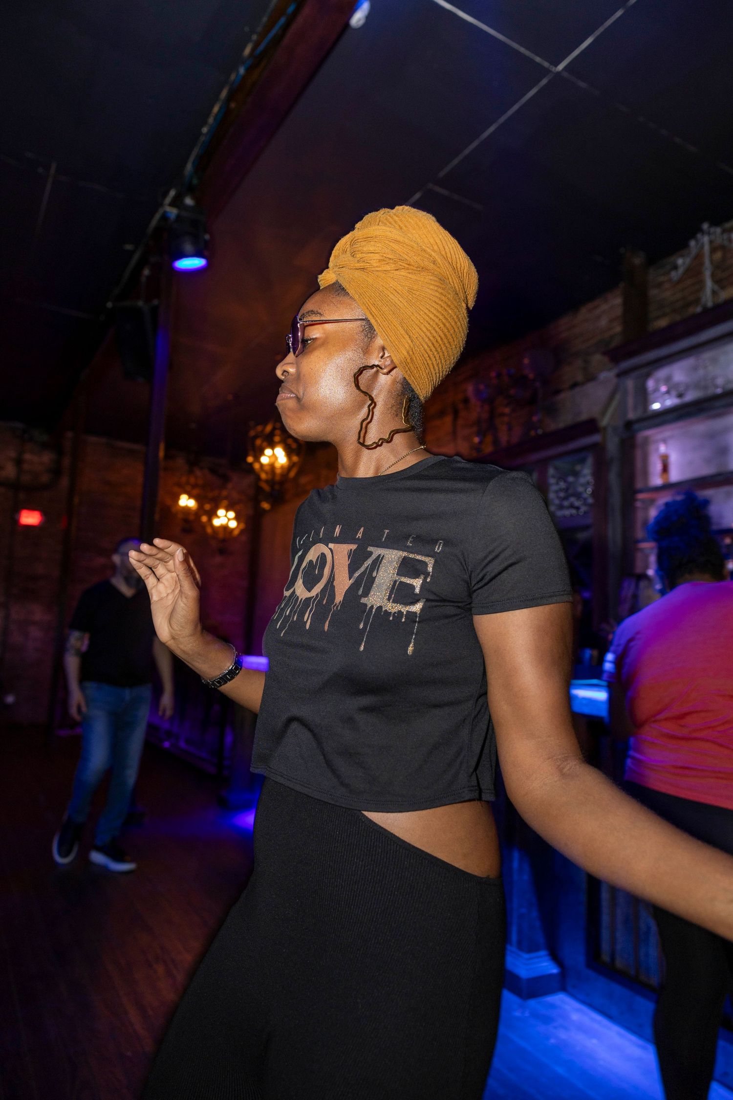 A performer in a yellow headwrap and 'LOVE' t-shirt dances at a dimly lit venue.