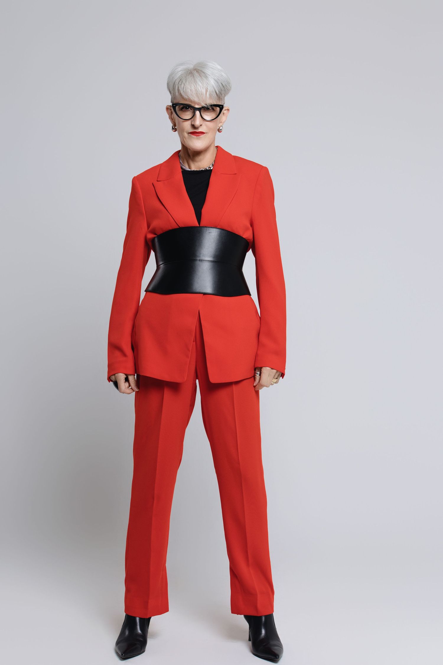 Stylish figure in bright red pantsuit with black corset belt and glasses strikes confident pose.