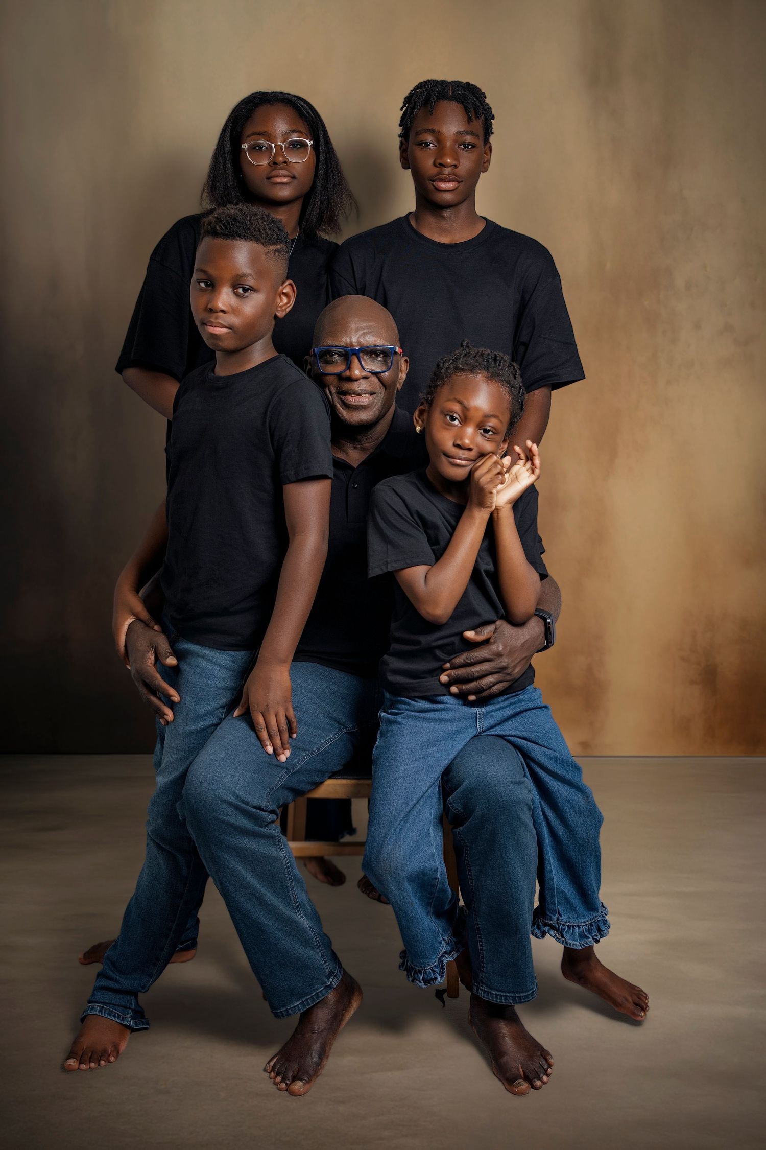 Two children wearing black shirts and jeans sit barefoot on a wooden surface against a textured brown backdrop.