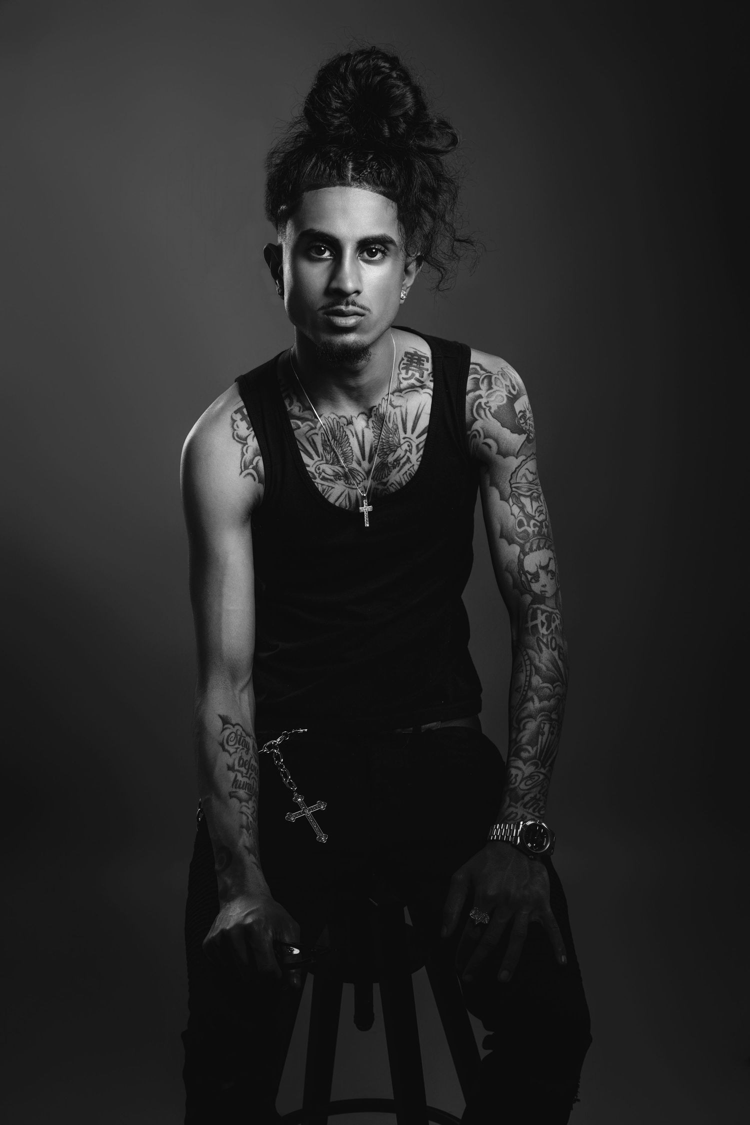 Black and white portrait of a tattooed person in a tank top seated on a stool.