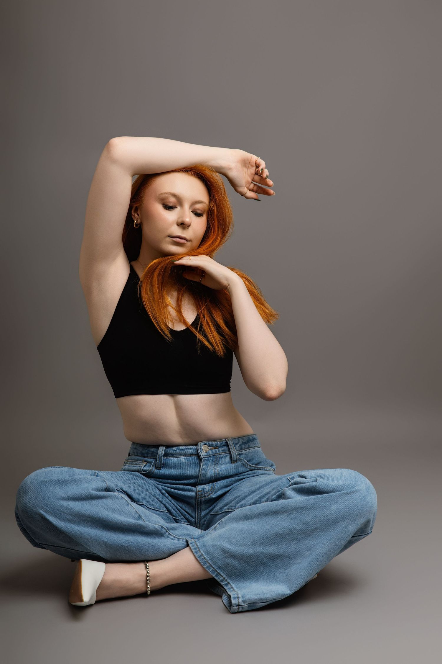 Artistic pose in black crop top and loose denim jeans on grey background.