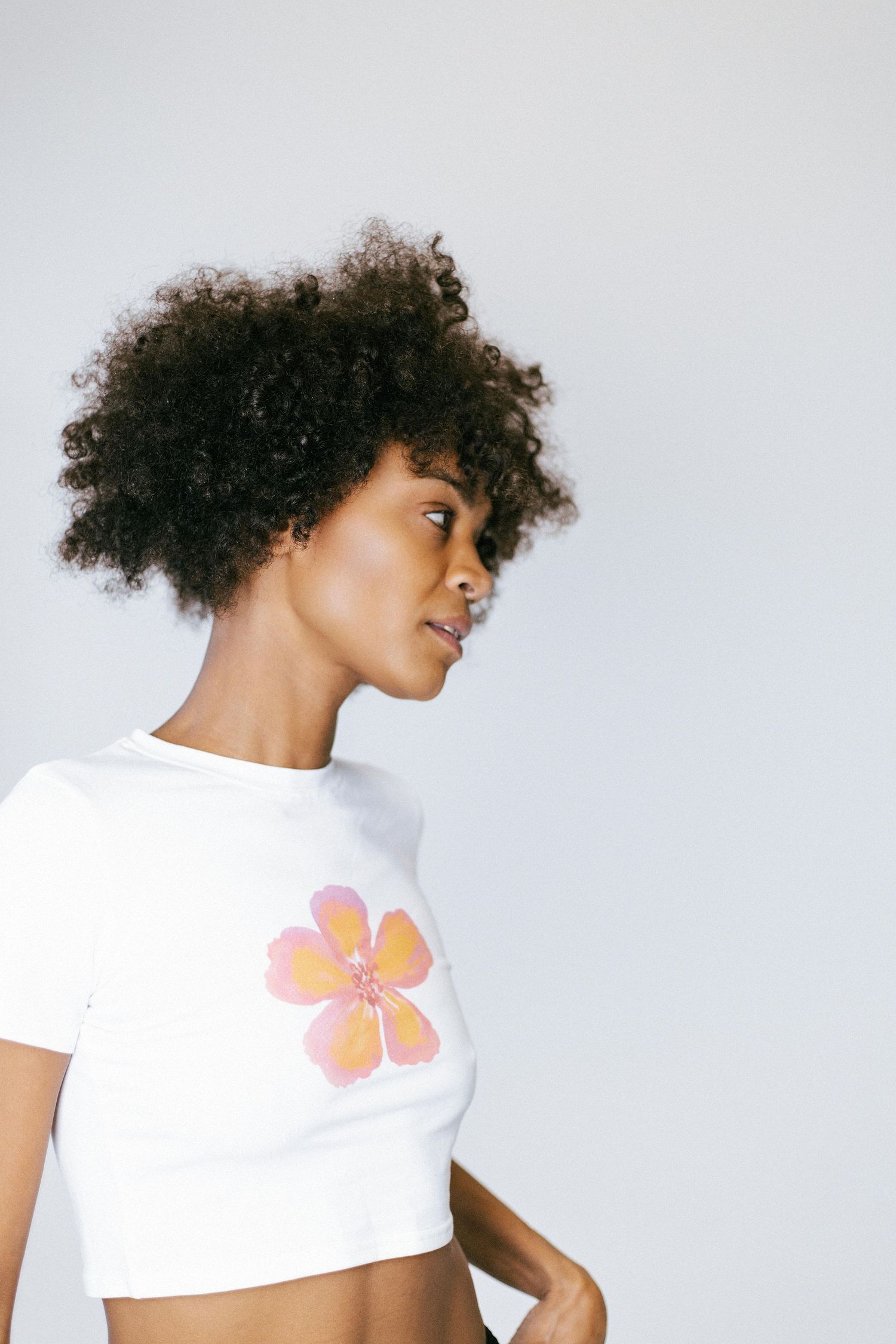 Person with curly hair wearing white t-shirt with pink flower graphic shown in multiple cropped poses against light background.