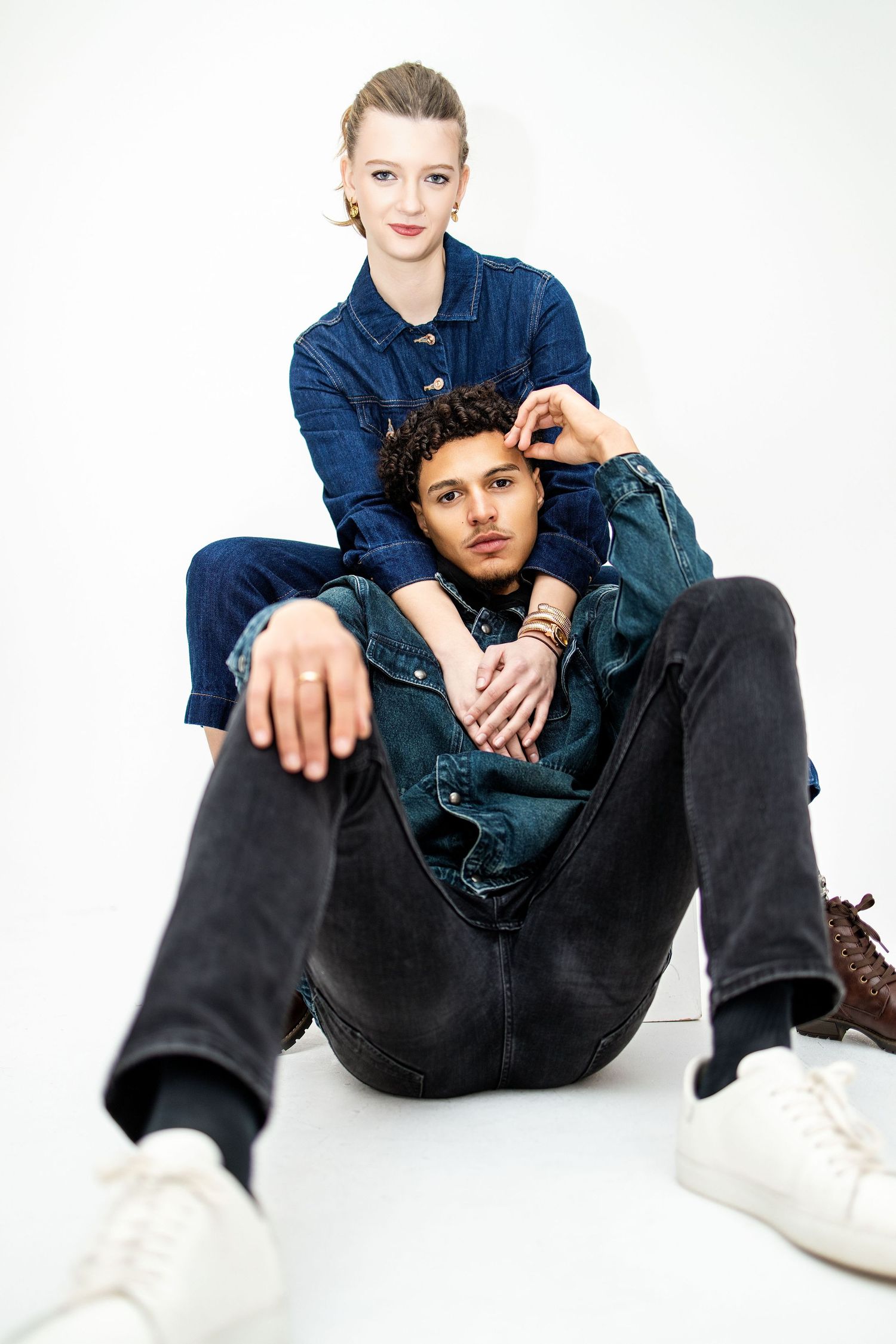 Person wearing denim jacket and black pants sits casually on white background in fashion editorial style pose.