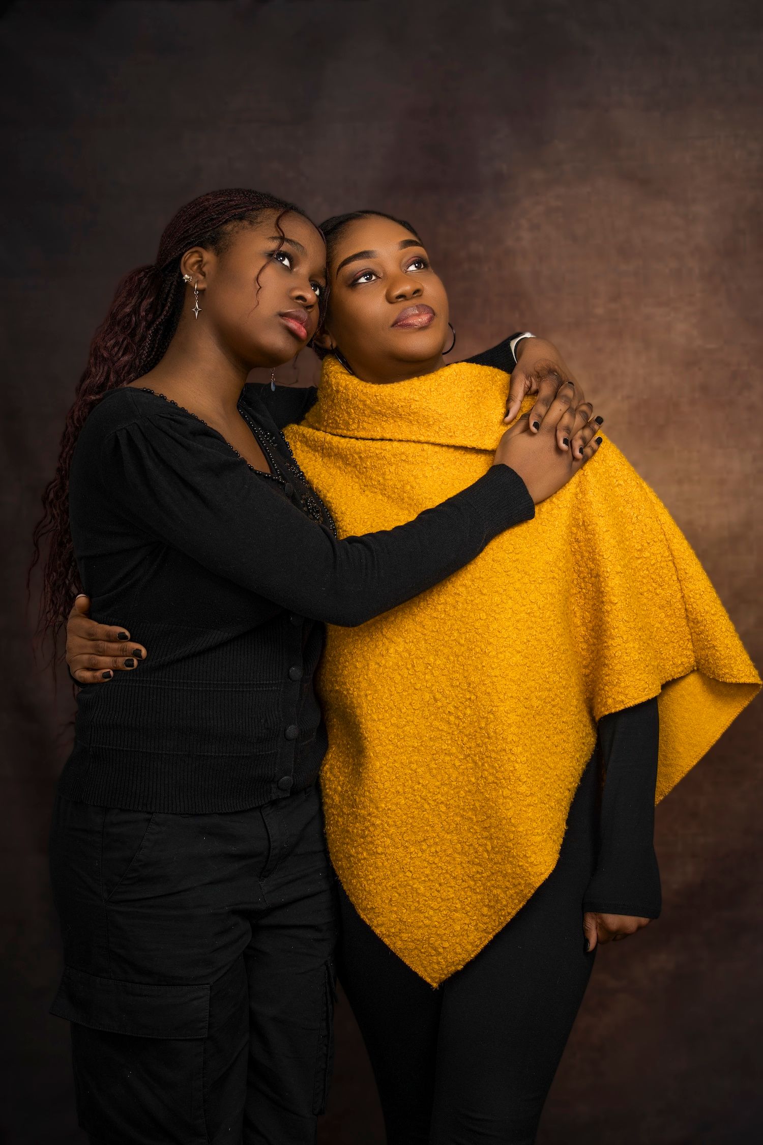 Two people embrace while wearing contrasting black and bright yellow clothing against a dark brown backdrop.
