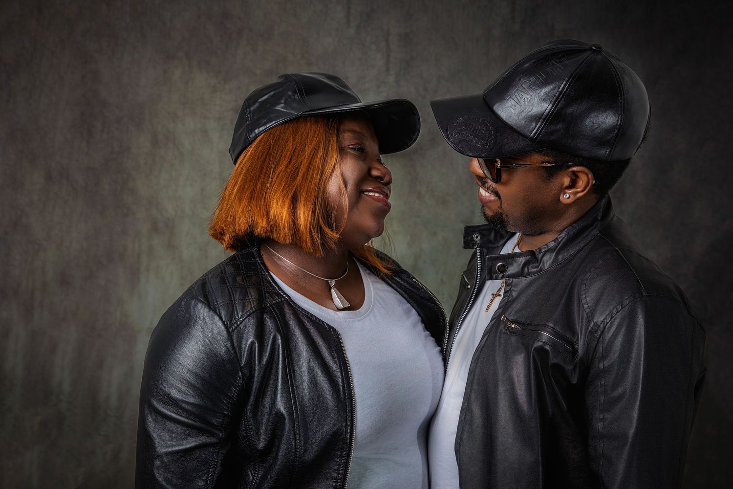 A couple in leather jackets and casual wear poses intimately against a dark backdrop.