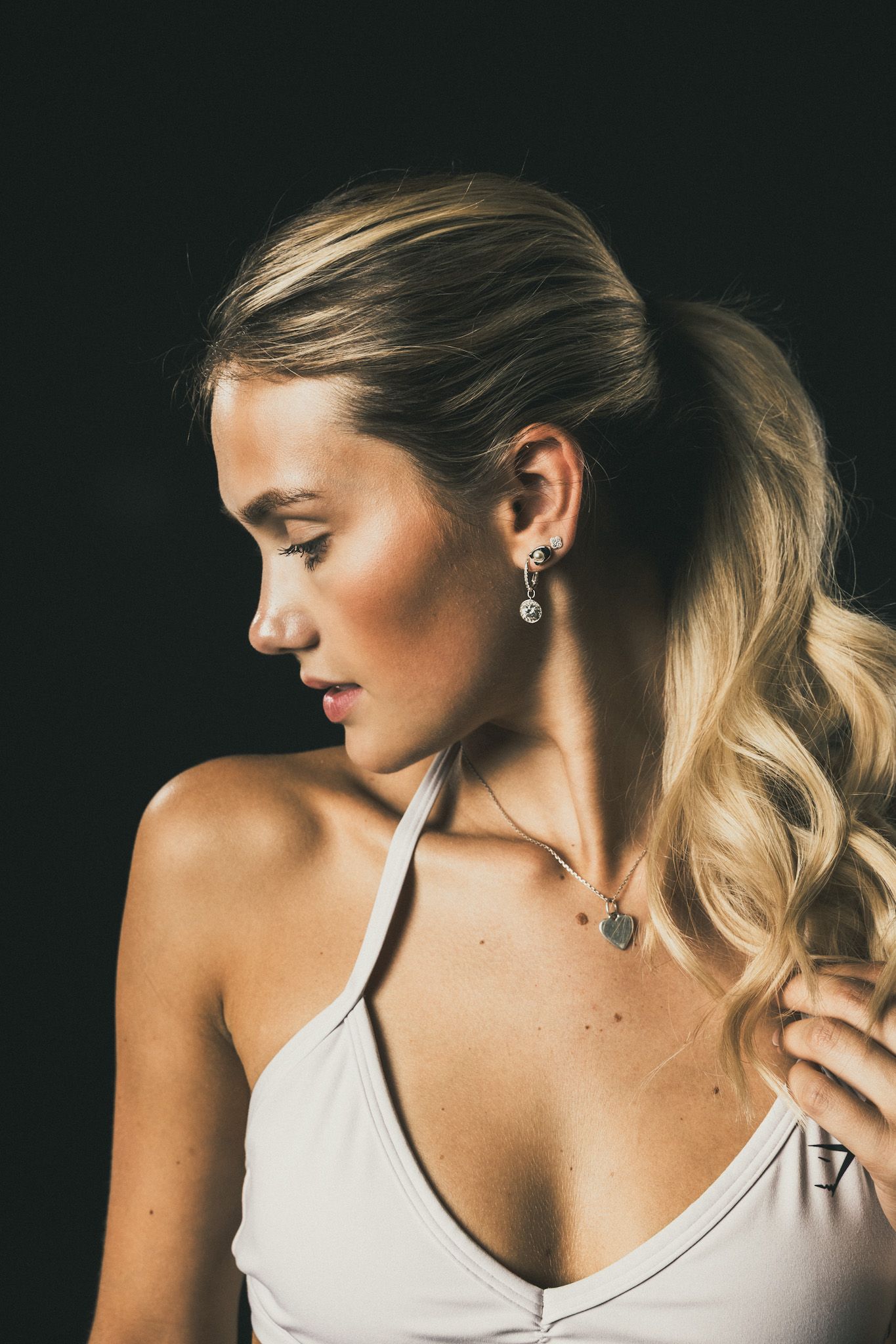 Blonde woman in white halter top with dangling earrings and necklace, posing against dark background.