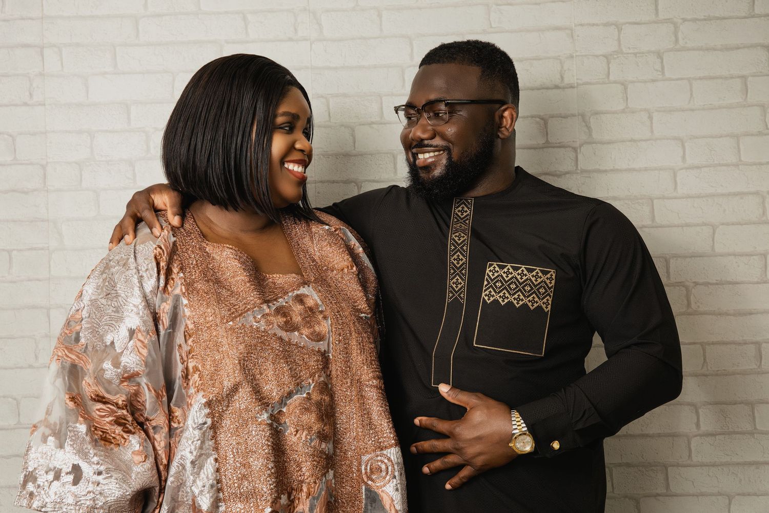 Couple sharing joyful moments against a white brick wall wearing elegant attire.