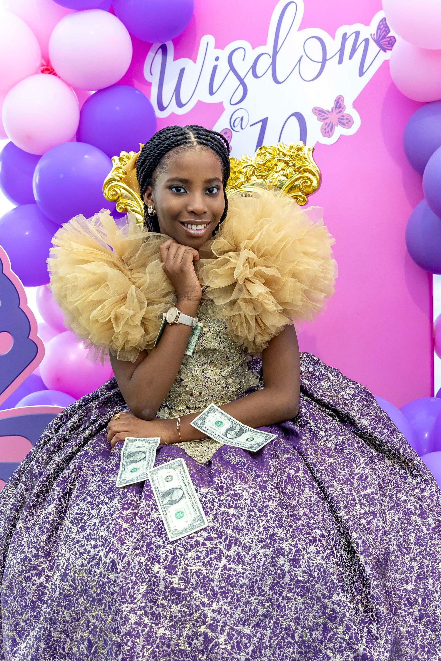 Young person in a sparkly purple and gold princess dress posing with money against pink balloon backdrop.