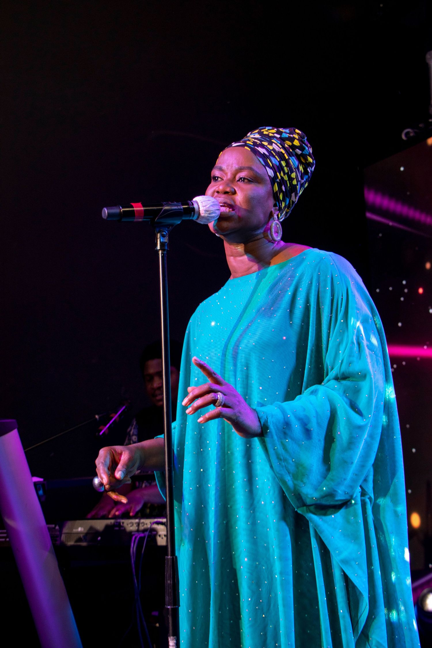 Performer in a sparkling turquoise gown and patterned headwrap singing on stage with dramatic lighting.