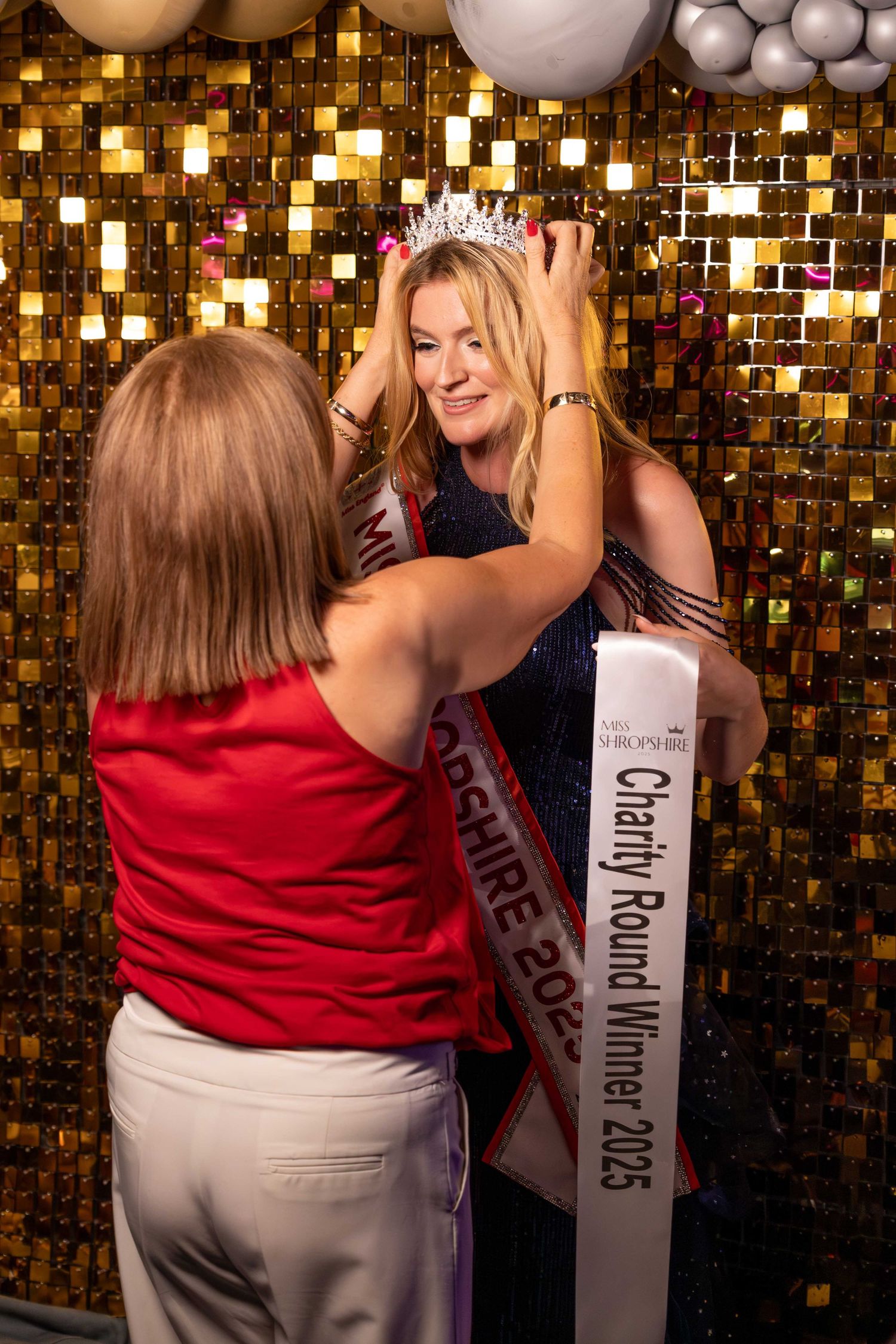 Women posing against a gold sequin backdrop with white balloons at a celebration event.