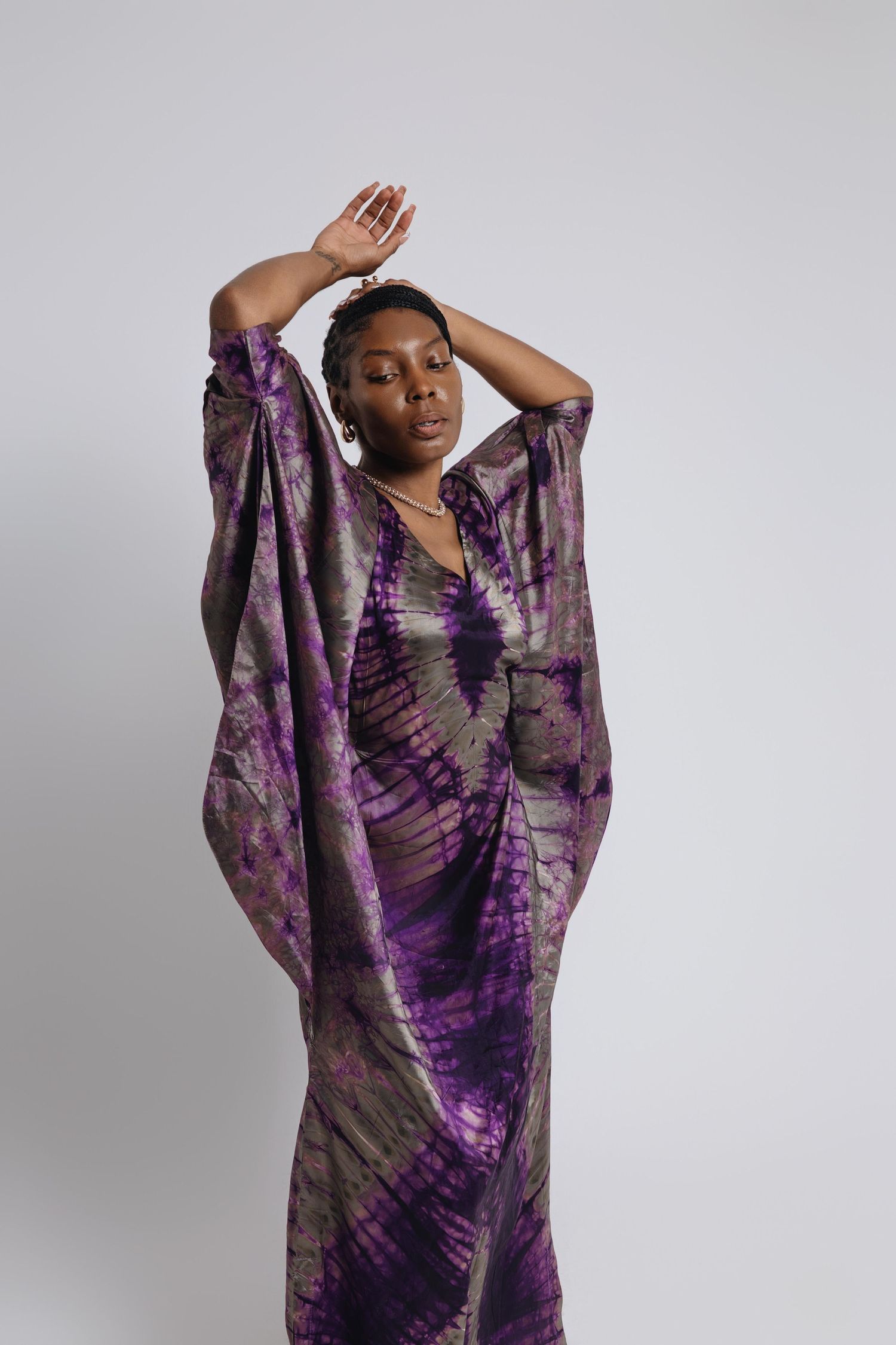 Model wearing flowing purple tie-dye dress poses gracefully against grey backdrop.