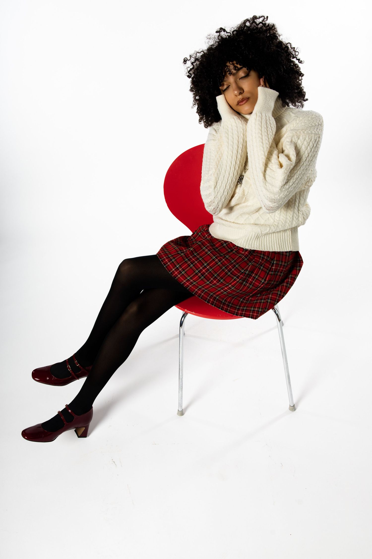Model in cozy white sweater and plaid skirt poses on white chair in winter fashion shoot.