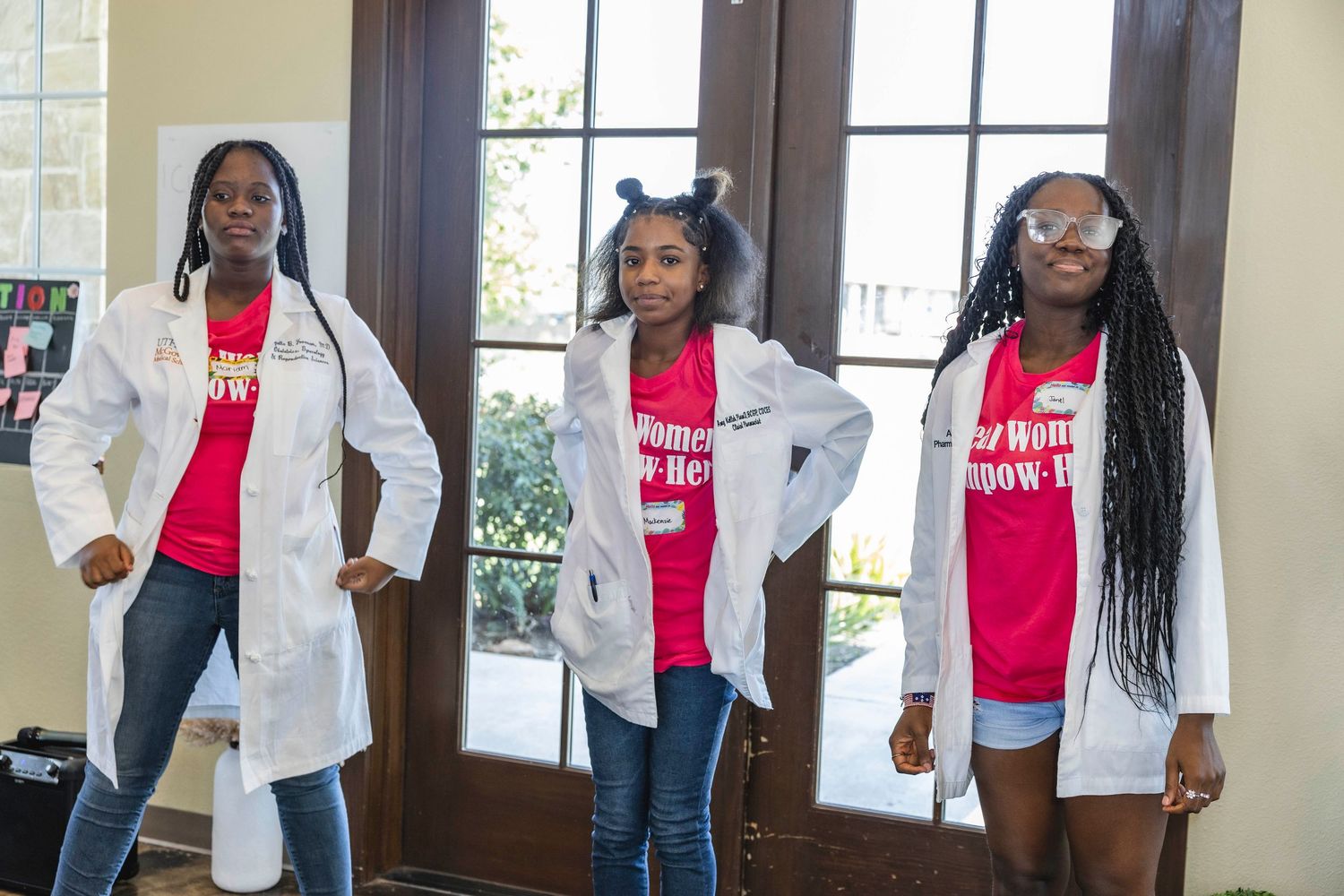Three people in white jackets and pink shirts posing confidently by windows.