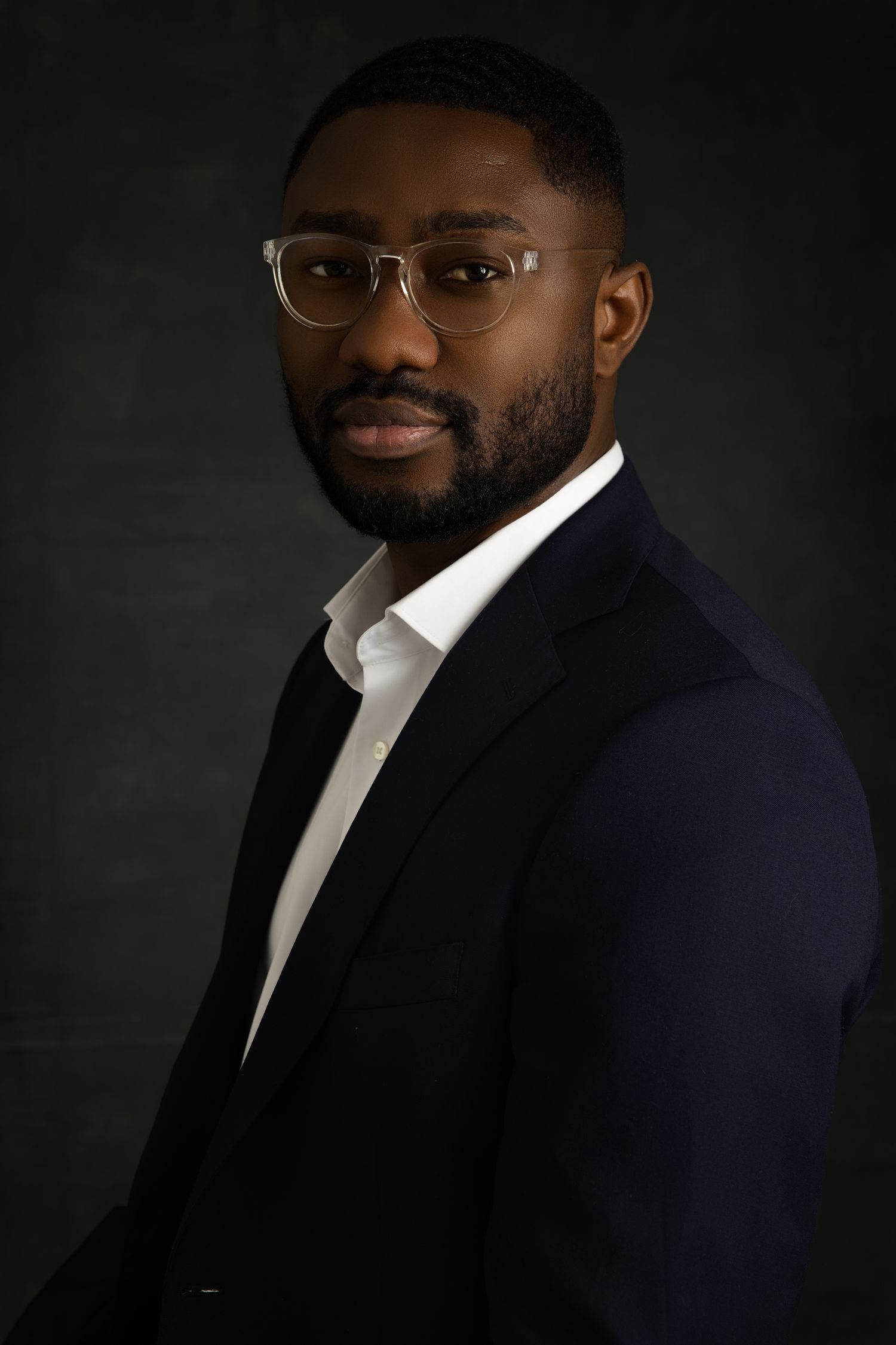 Professional portrait of someone in a navy suit jacket and white dress shirt against a dark background.