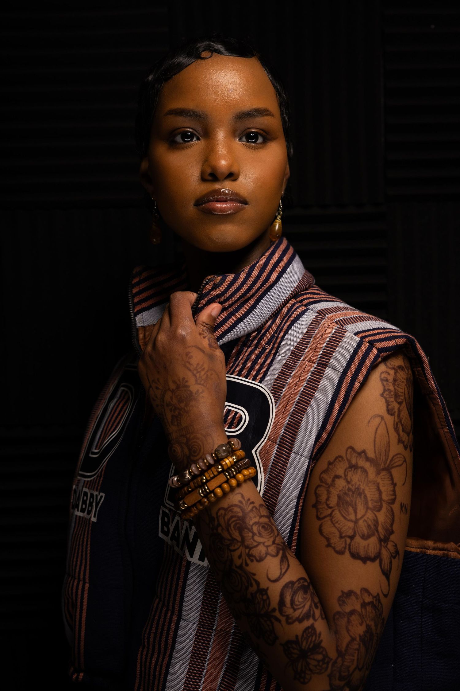 Artistic portrait showing intricate arm tattoos and striped collar in low-key lighting.
