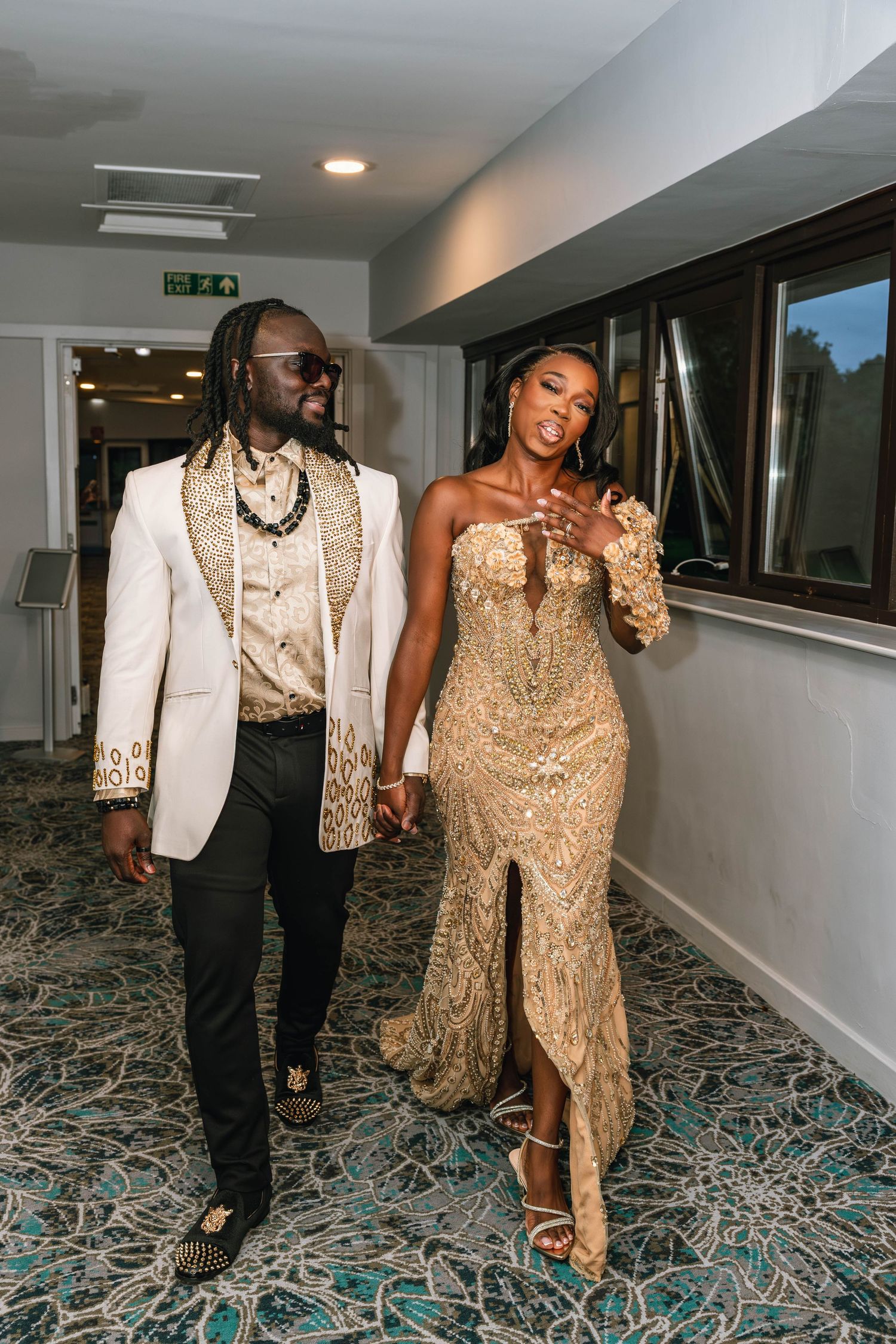 Wedding couple walking together in formal attire through a carpeted hallway.