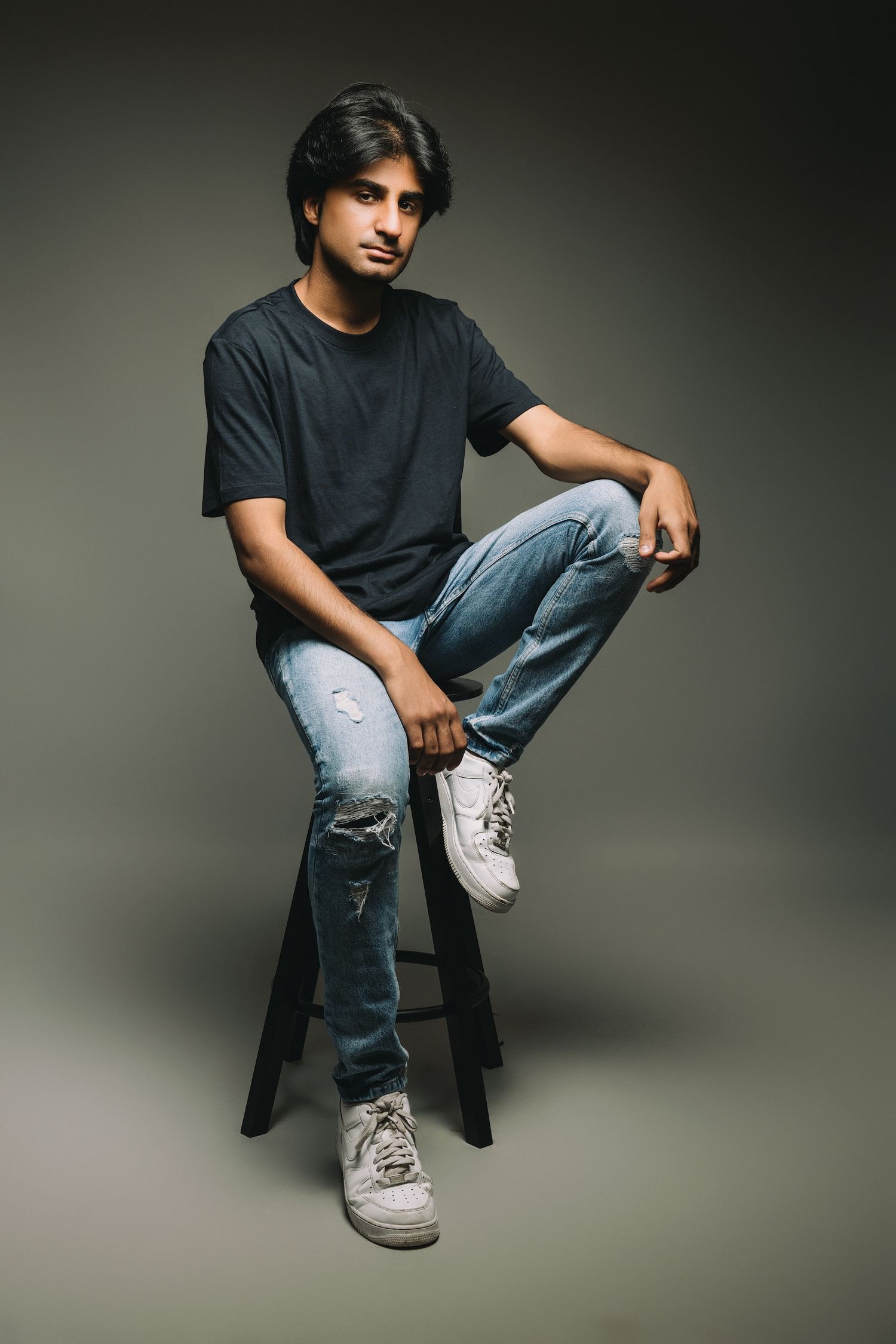 Person in casual black t-shirt and jeans sits on dark stool against gray backdrop in artistic portrait series.