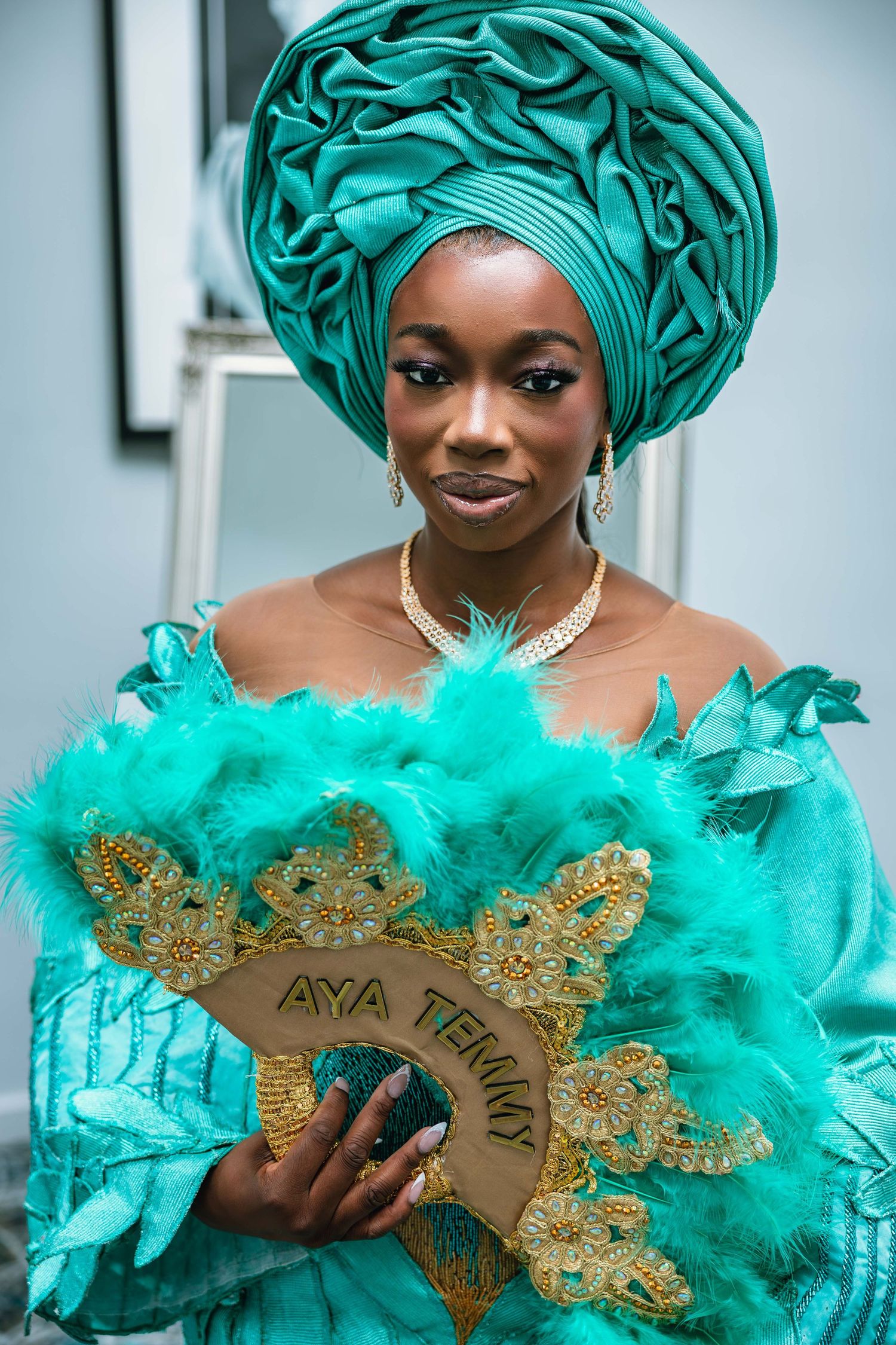 Traditional Nigerian fashion with turquoise gele headwrap and feathered ceremonial fan.