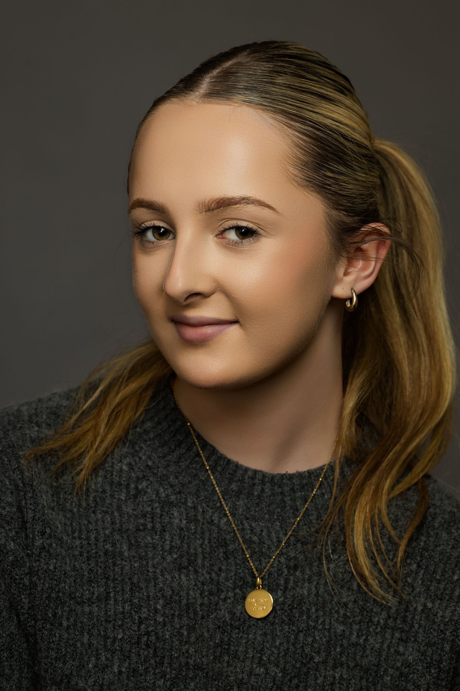 Studio portrait featuring a dark gray sweater and gold necklace against moody lighting.