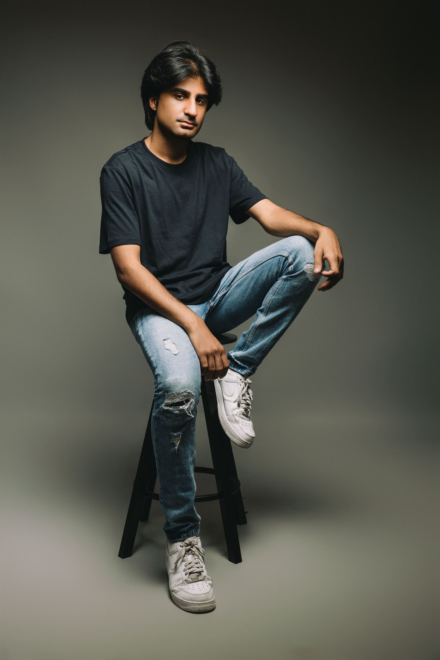 Studio portraits of someone in casual attire sitting on dark stool against gray background.