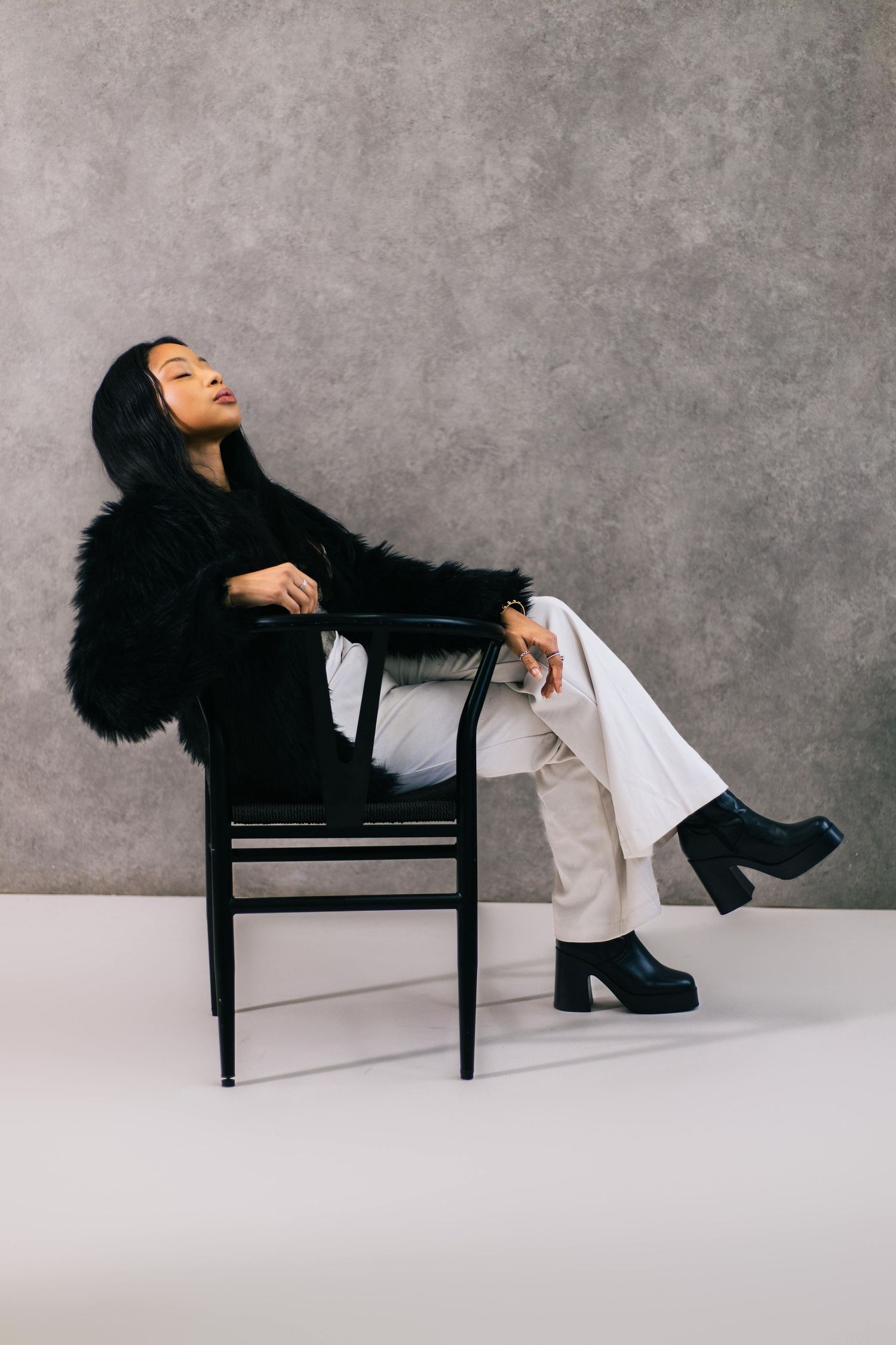Woman in black fur coat and white pants sitting on black chair against gray wall, wearing platform boots.