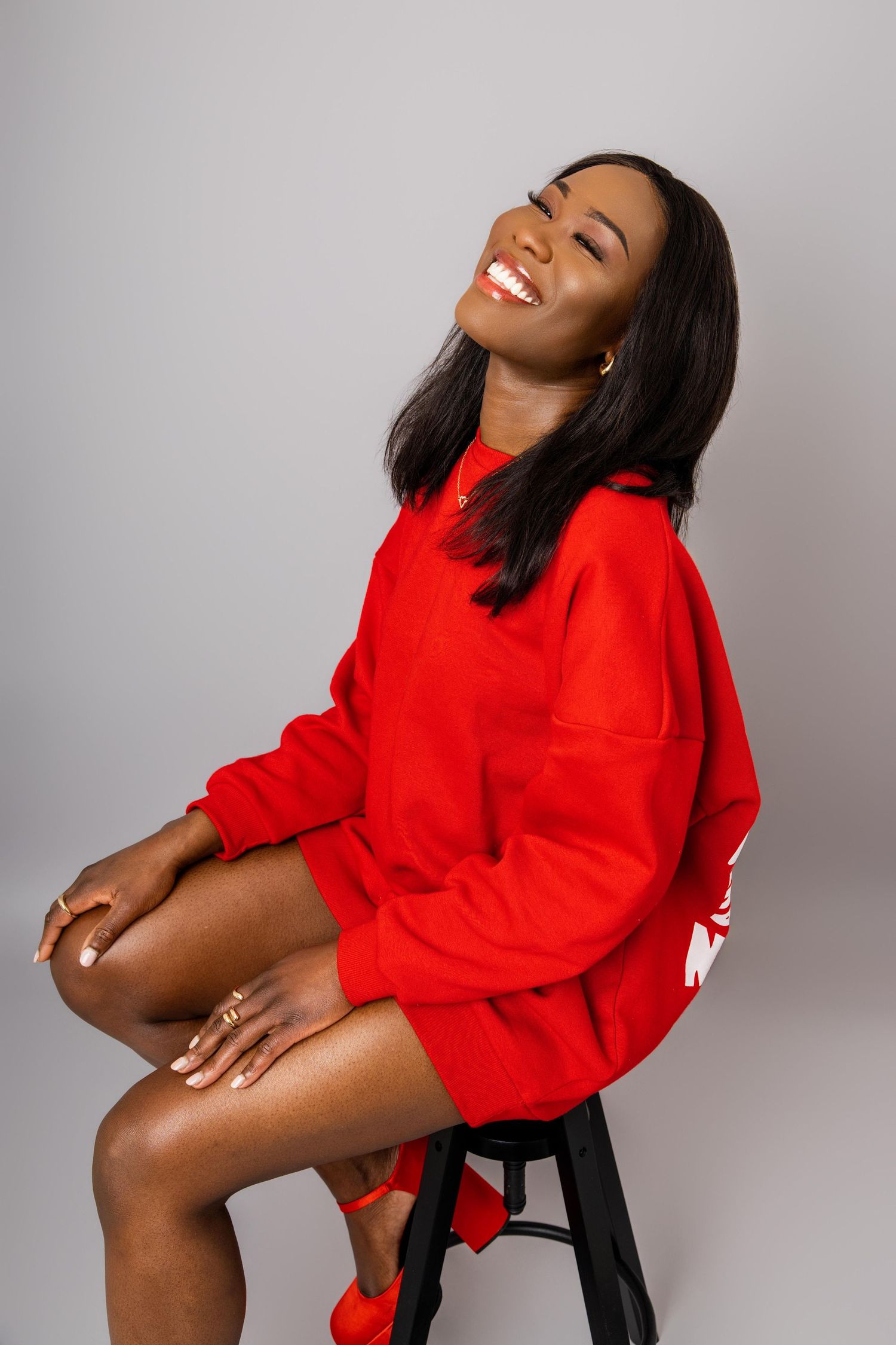 Model in vibrant red oversized sweater poses playfully on chair against light gray background.