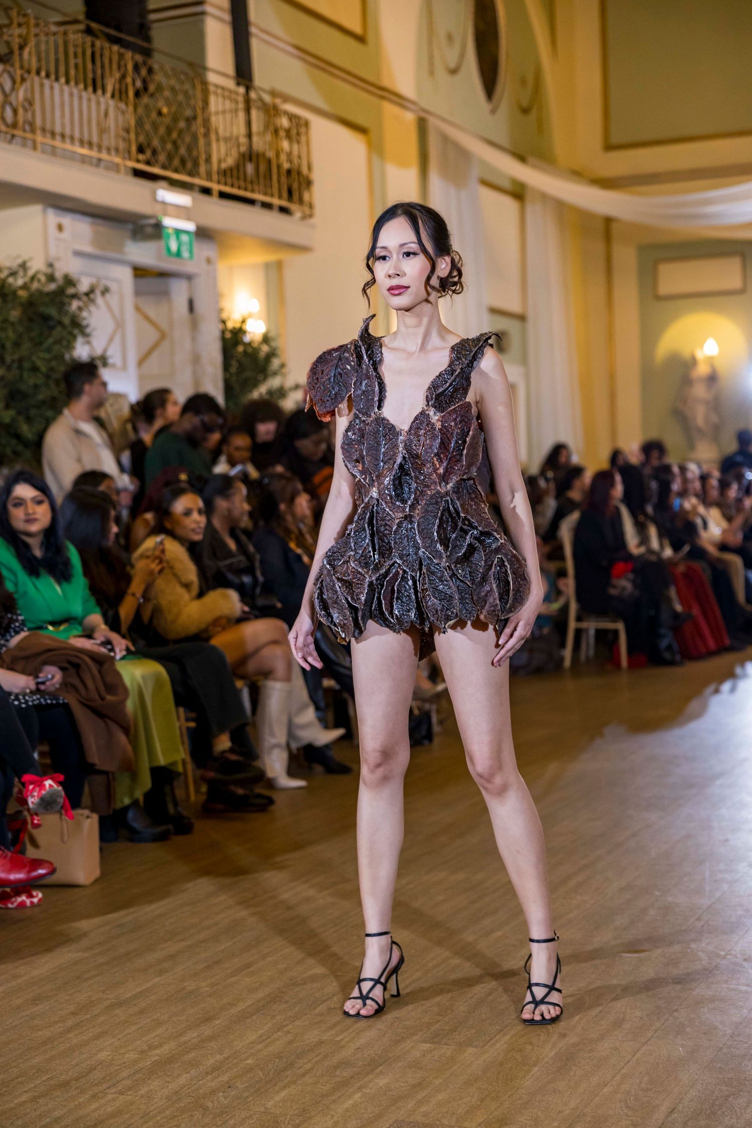 Models presenting short black patterned dresses on runway with elegant venue backdrop.