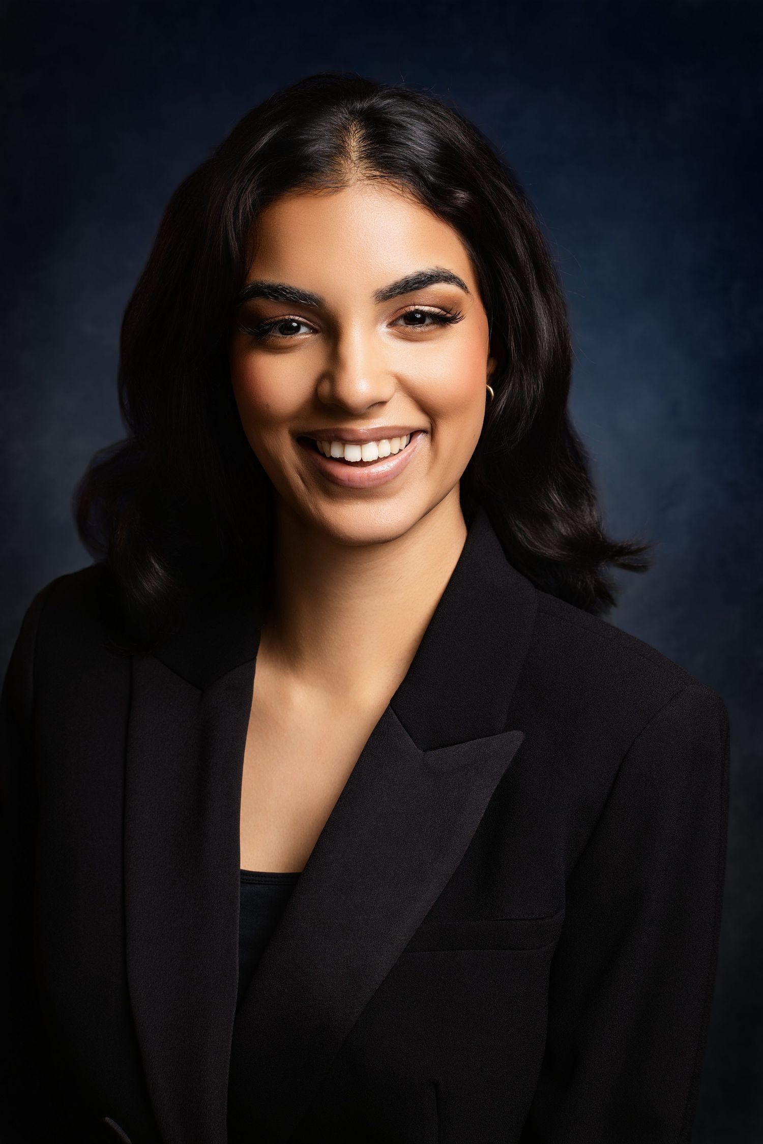Professional headshot of person in black blazer with wavy hair against dark background.