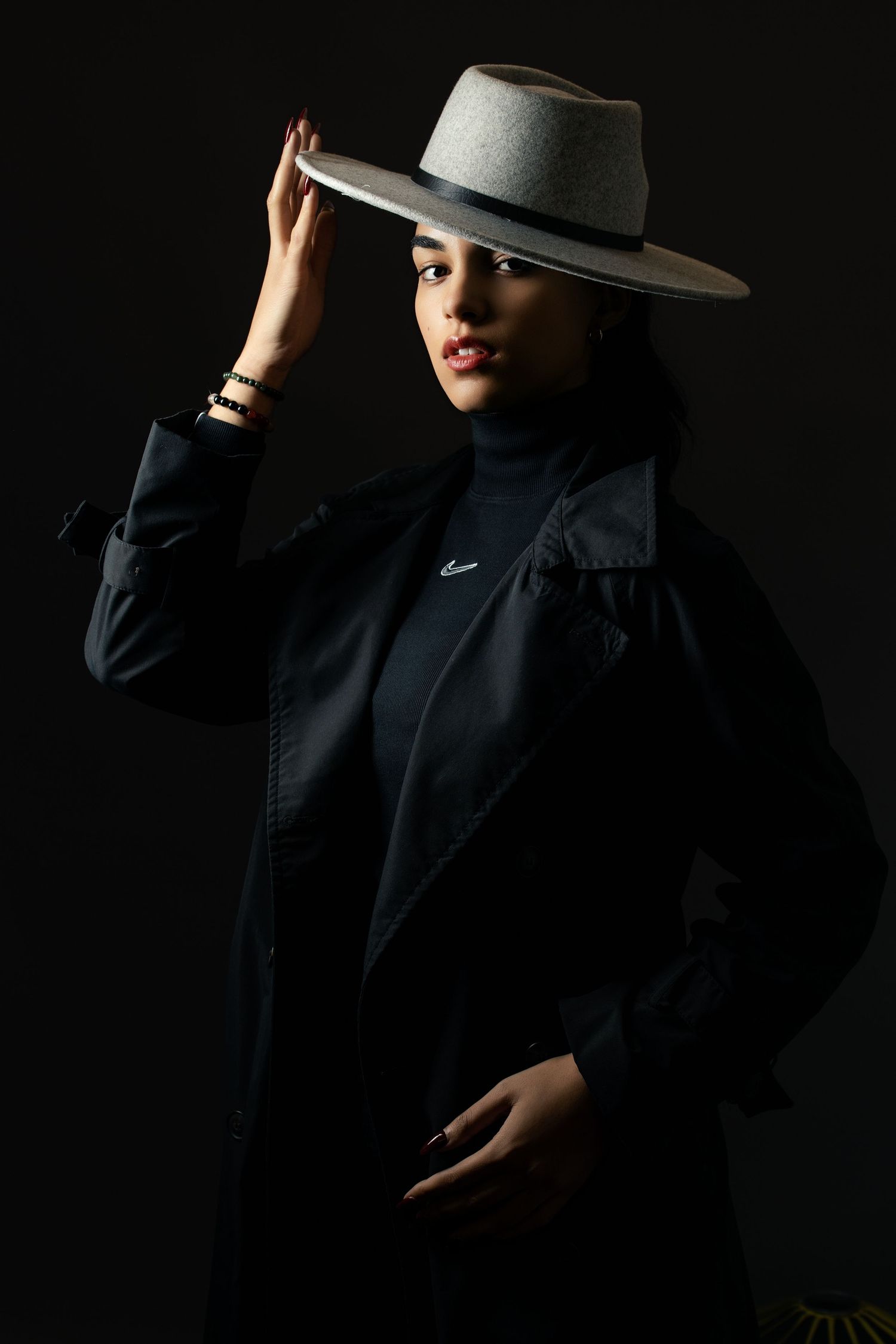 A stylish woman in a black jacket and turtleneck poses confidently, holding a wide-brim gray felt hat against a dark background.