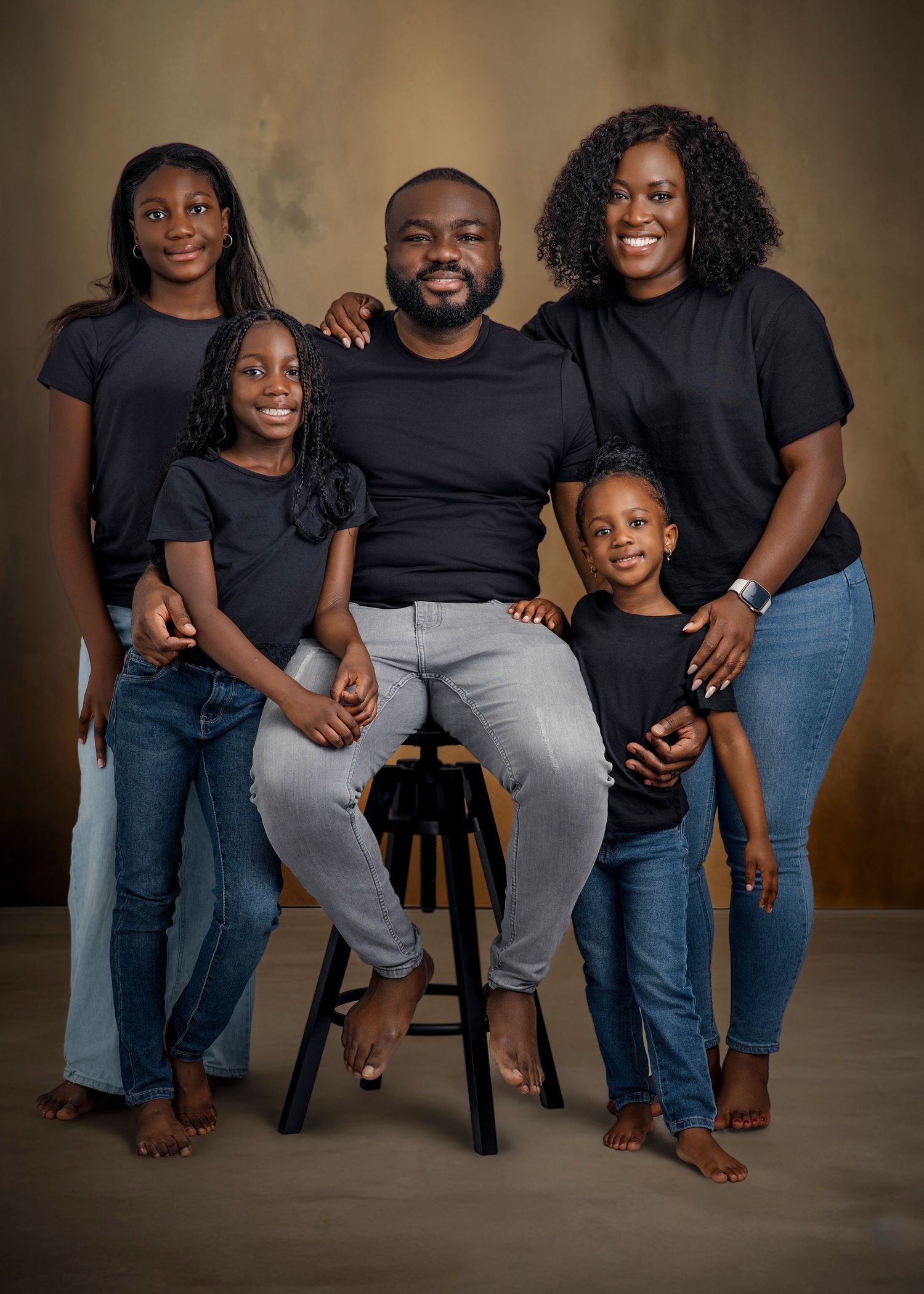 Family wearing coordinated black outfits poses together in studio portraits against warm brown backdrop.