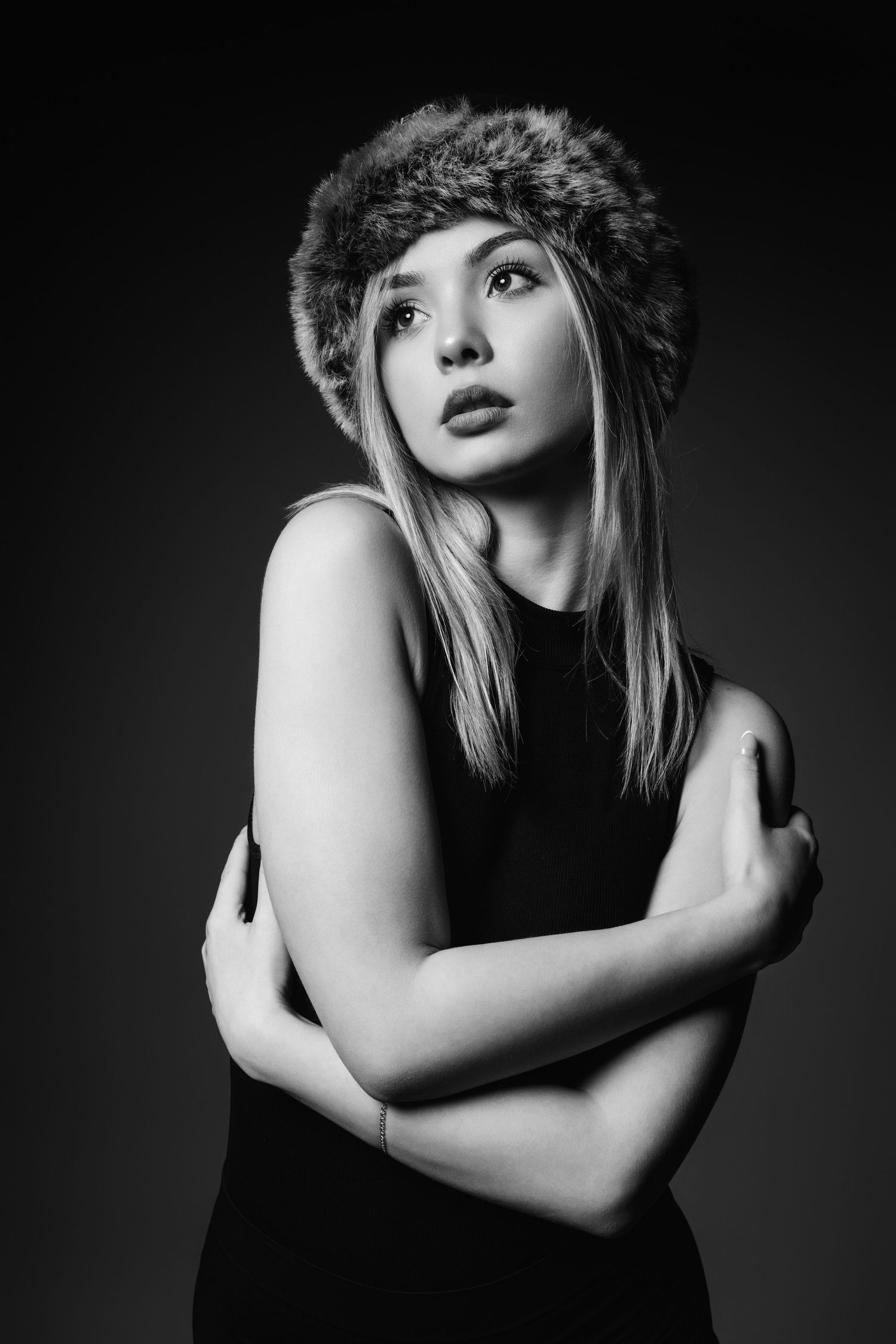 Dramatic black and white portrait of a person wearing a fur hat and sleeveless top against dark background.