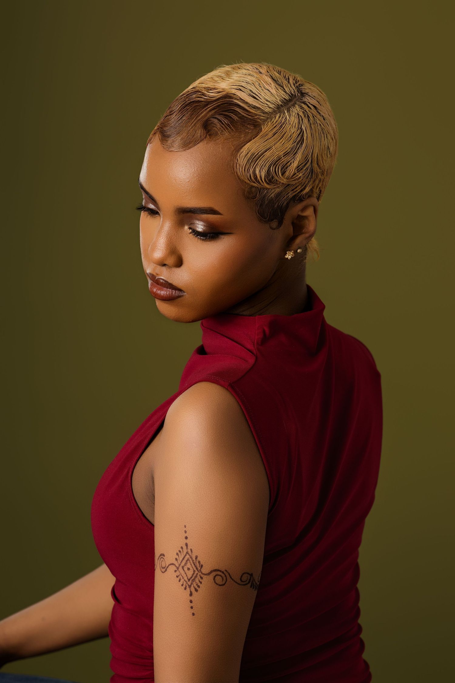 Artistic portrait of a woman in red turtleneck with short blonde hair against olive background.