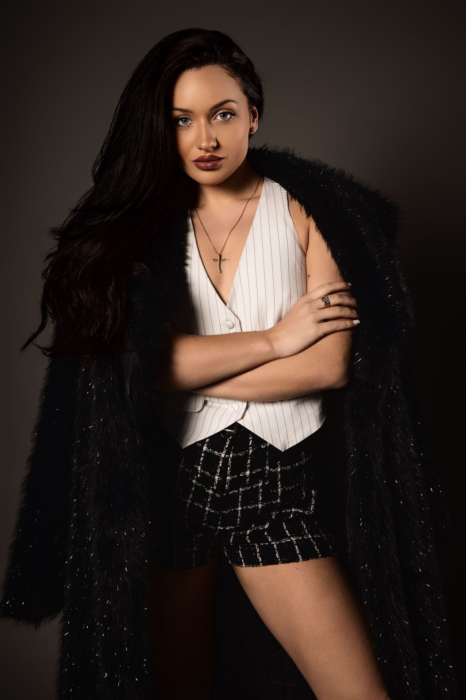Woman in black fur coat, white pinstripe vest, and plaid shorts poses with arms crossed in studio lighting.