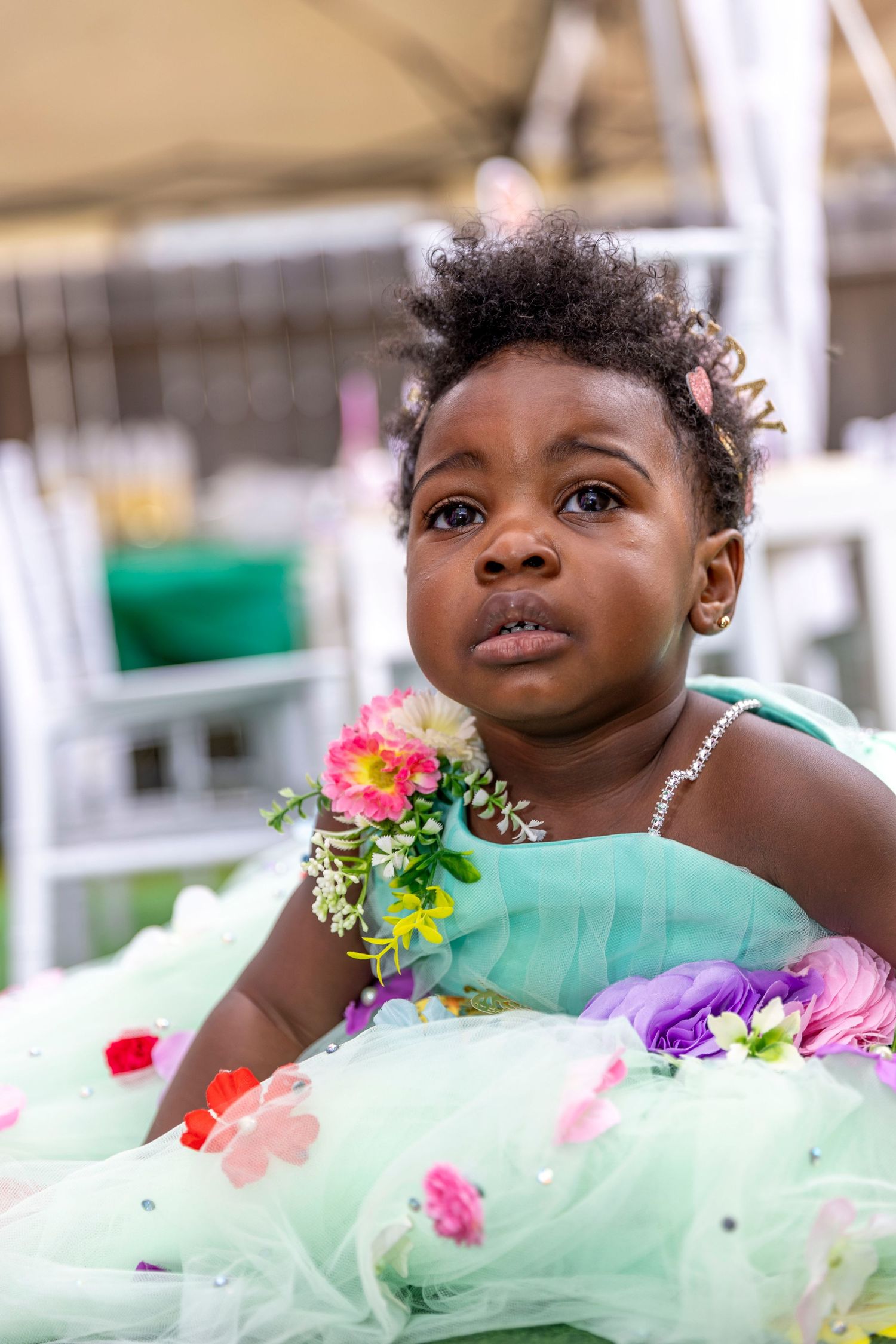Young child in a mint green dress adorned with colorful floral decorations poses for birthday photos.
