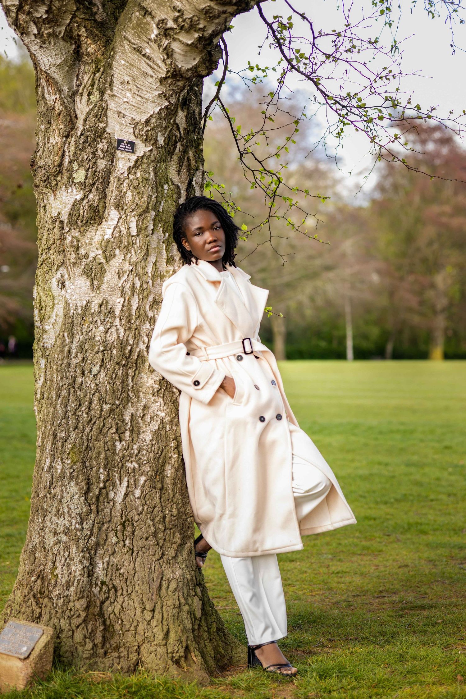 Elegant cream coat ensemble photographed against spring trees in park setting.