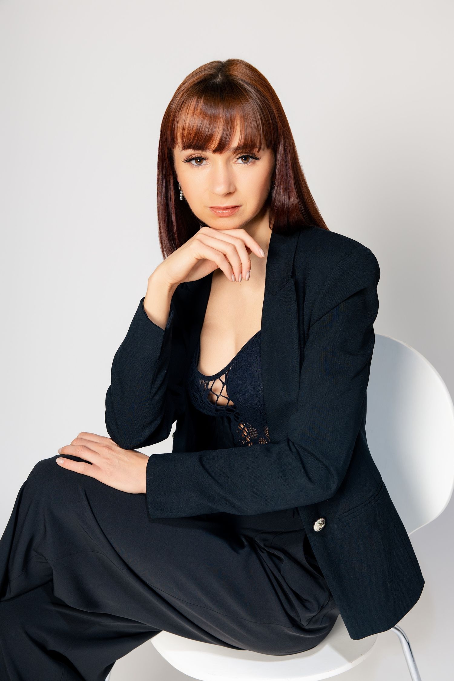 Woman in black blazer poses thoughtfully on white chair in professional studio photoshoot series.