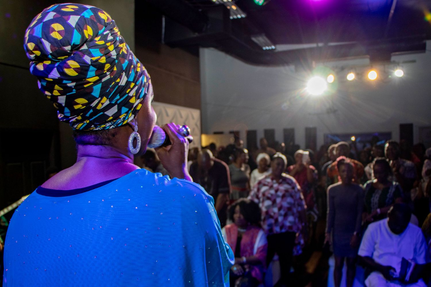 Performer in blue shirt and colorful headwrap faces crowd at nighttime event with stage lighting.