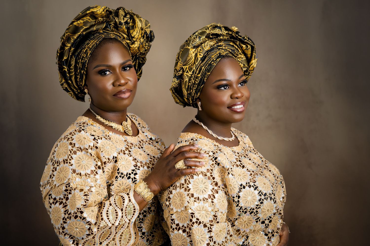 Two people wearing matching gold patterned outfits and head wraps pose together in a studio portrait.