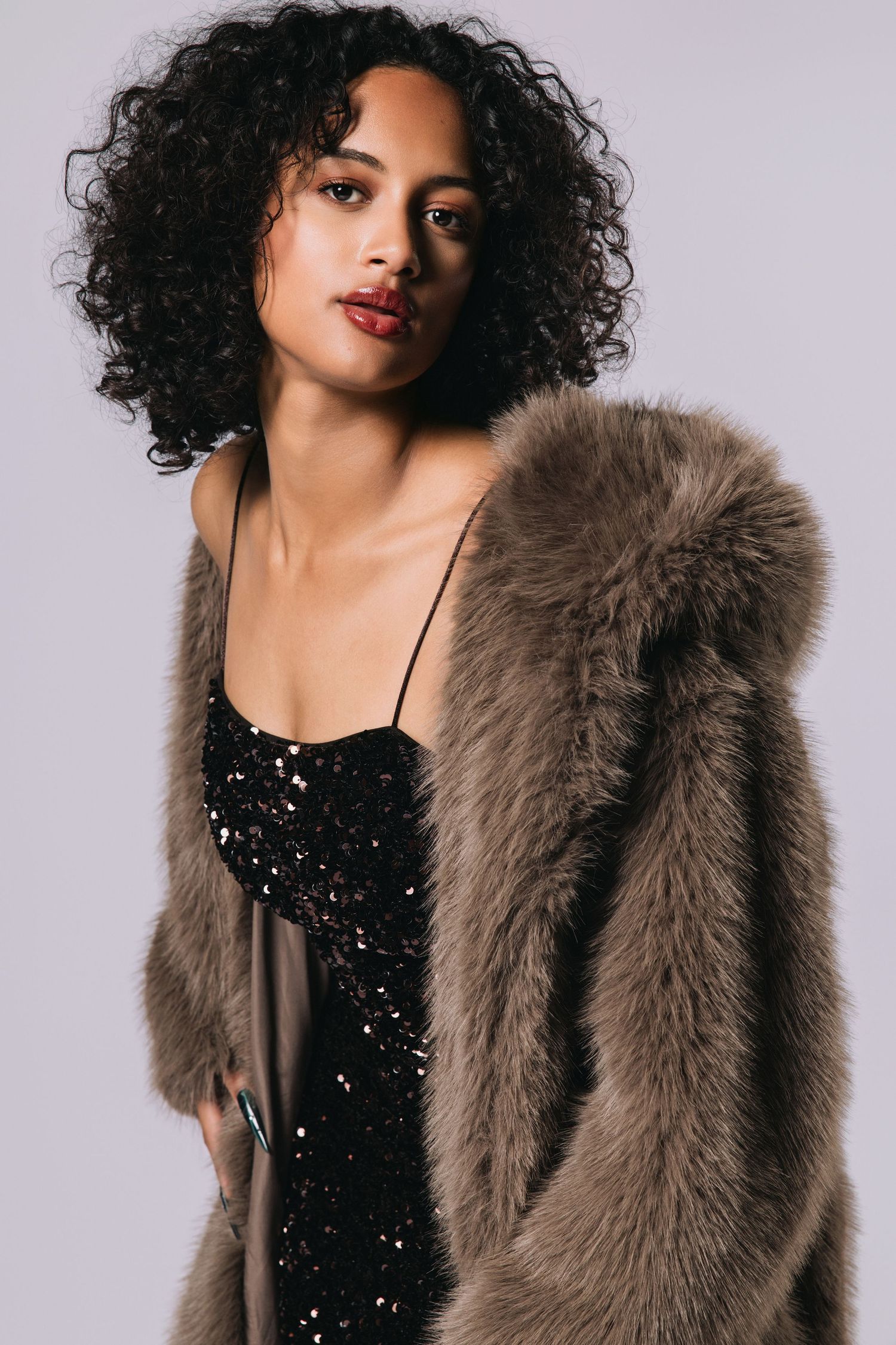 Woman in black sequin dress and taupe faux fur coat posing against gray background.