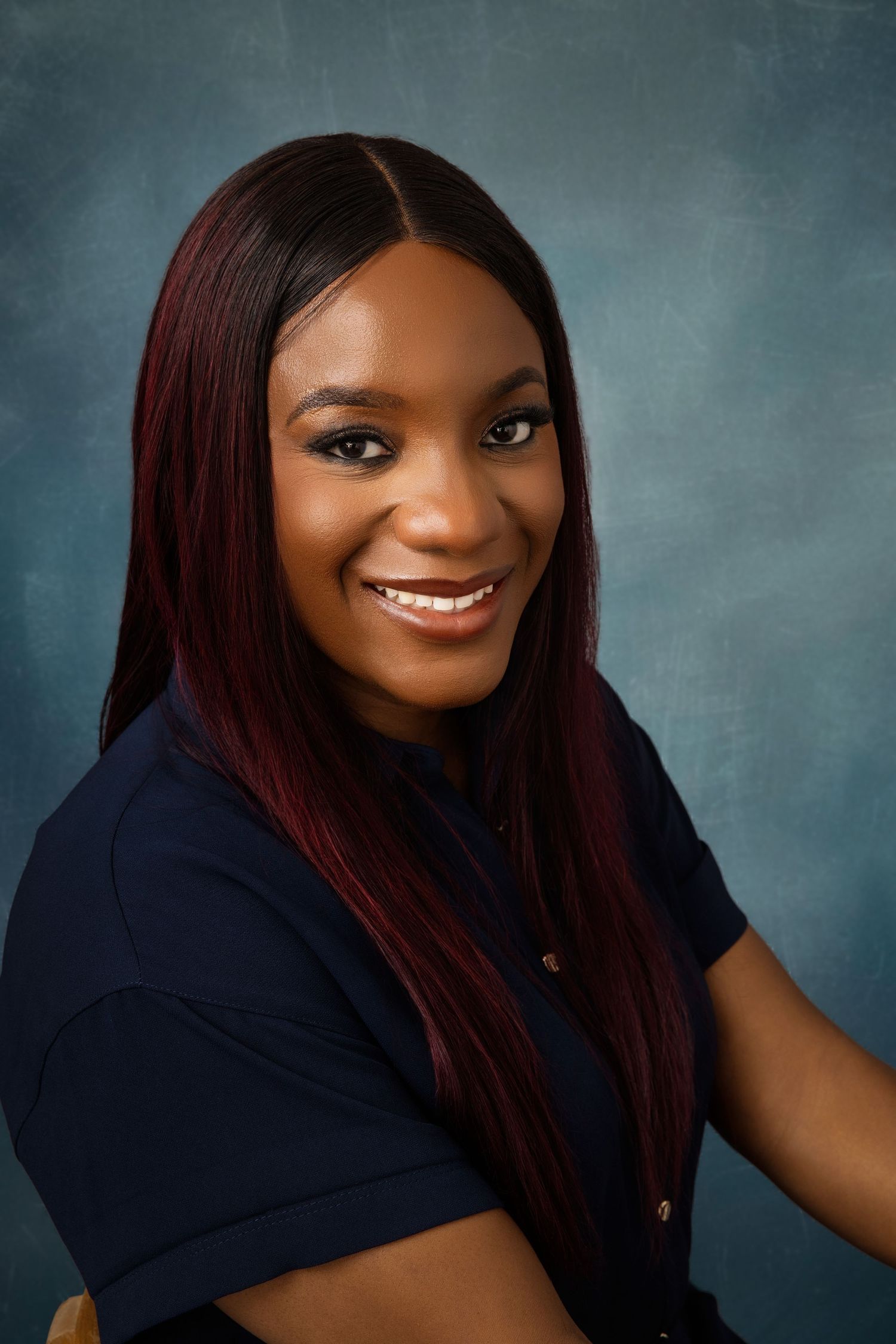 Professional headshot featuring burgundy-tinted hair and navy blue top against teal background.